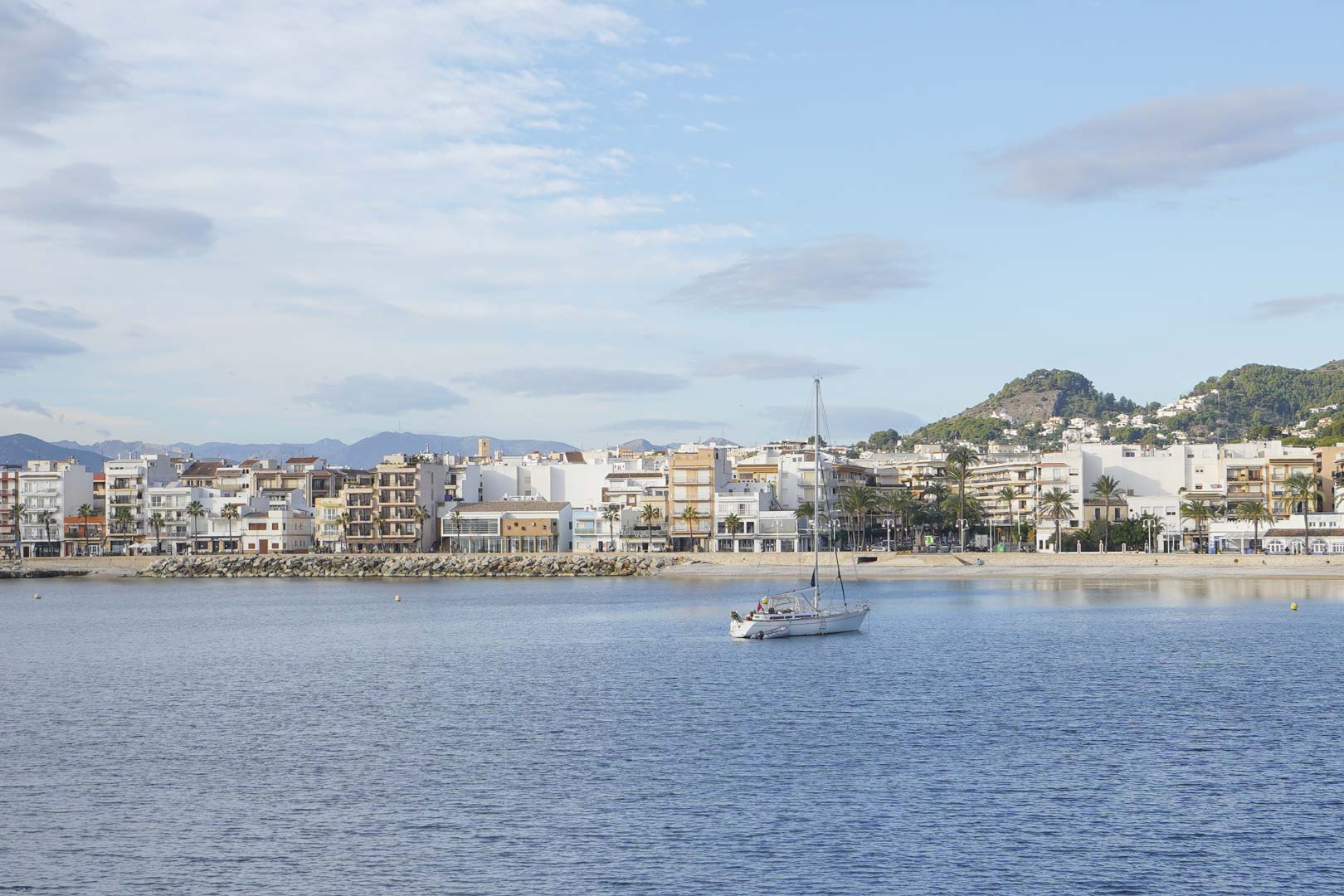 Vue panoramique sur le port de Jávea avec un voilier au mouillage, bordée par des bâtiments méditerranéens et des collines verdoyantes.
