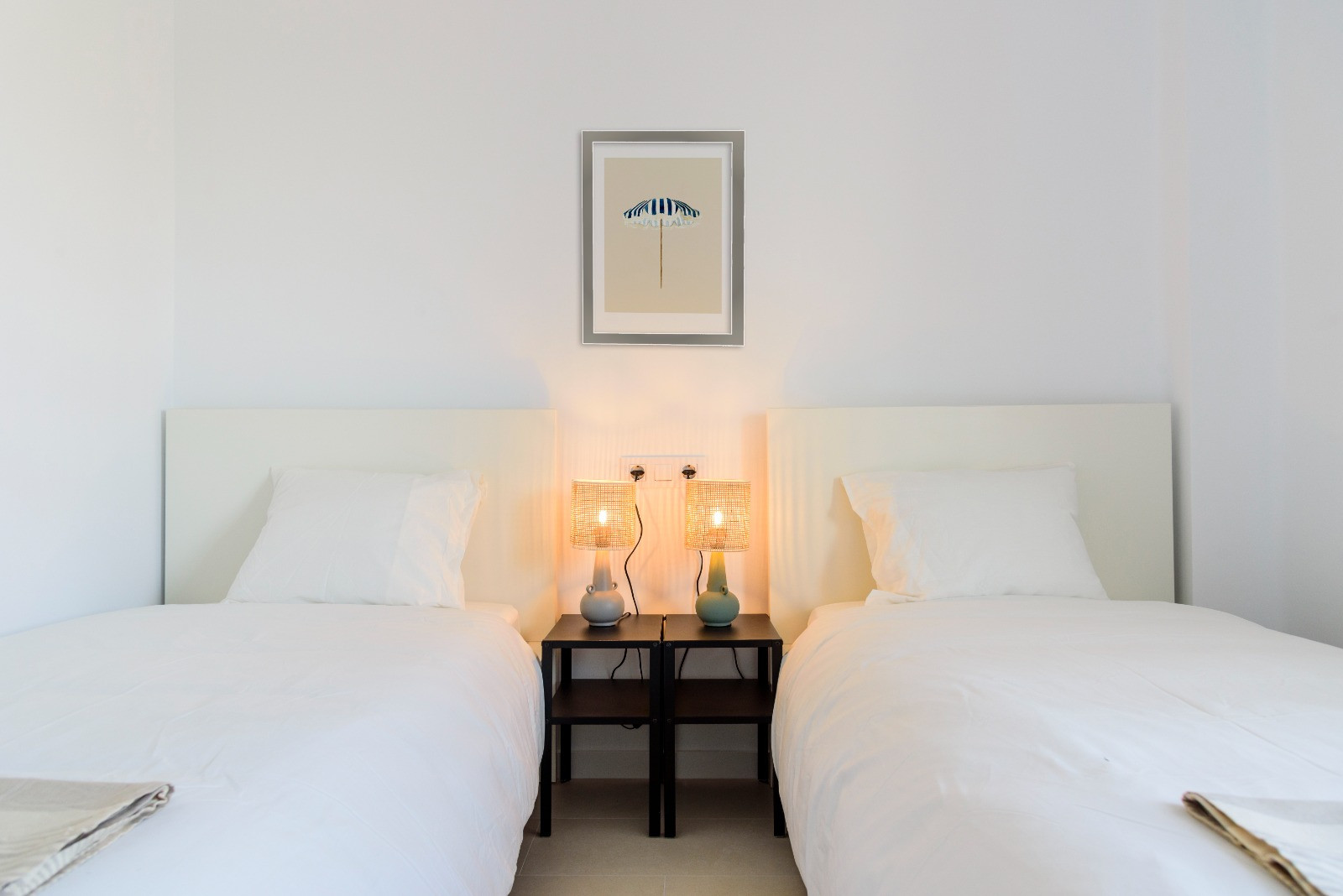 Chambre à deux lits simples avec têtes de lit blanches, tables de chevet noires et lampes en rotin créant une atmosphère chaleureuse et épurée.