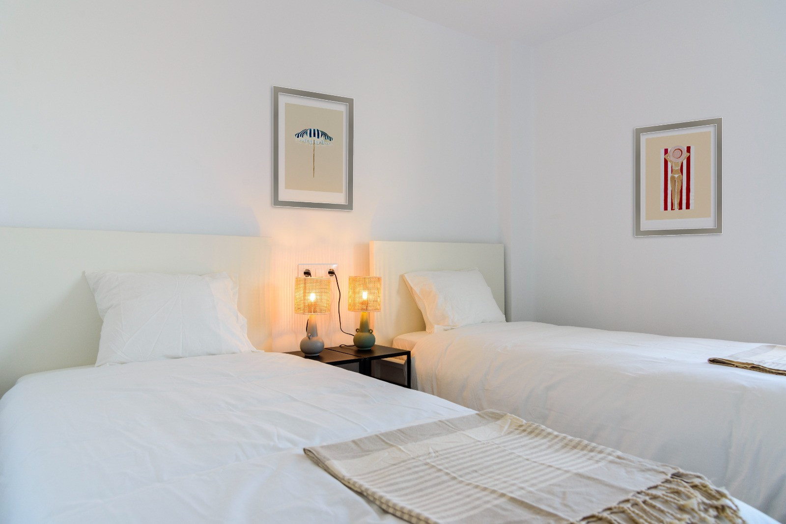 Chambre lumineuse avec deux lits simples, têtes de lit blanches et lampes en fibres naturelles. Murs blancs décorés de cadres aux motifs balnéaires épurés.