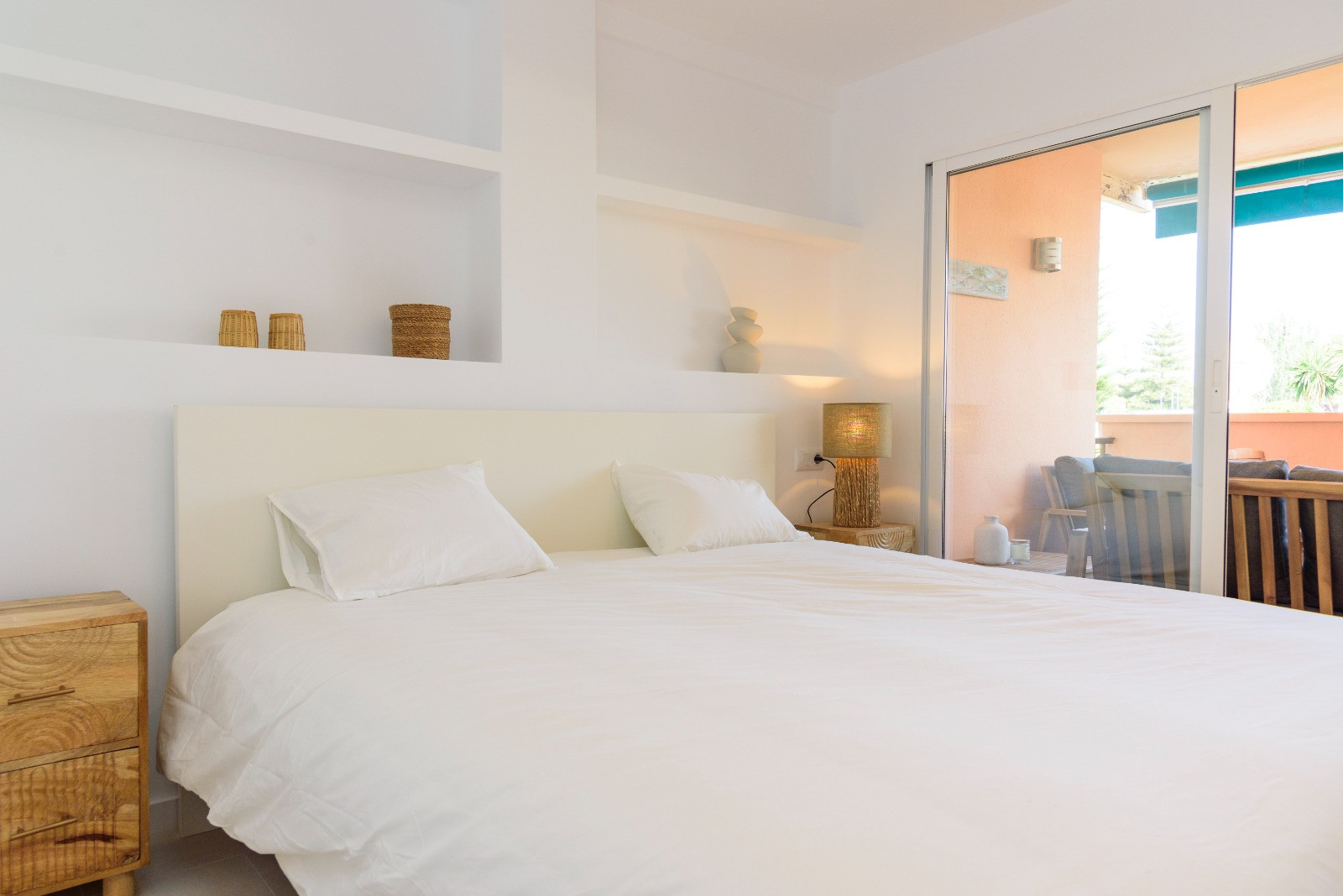 Chambre lumineuse avec étagères murales intégrées, tables de chevet en bois et accès direct à une terrasse privée par de grandes baies vitrées coulissantes.
