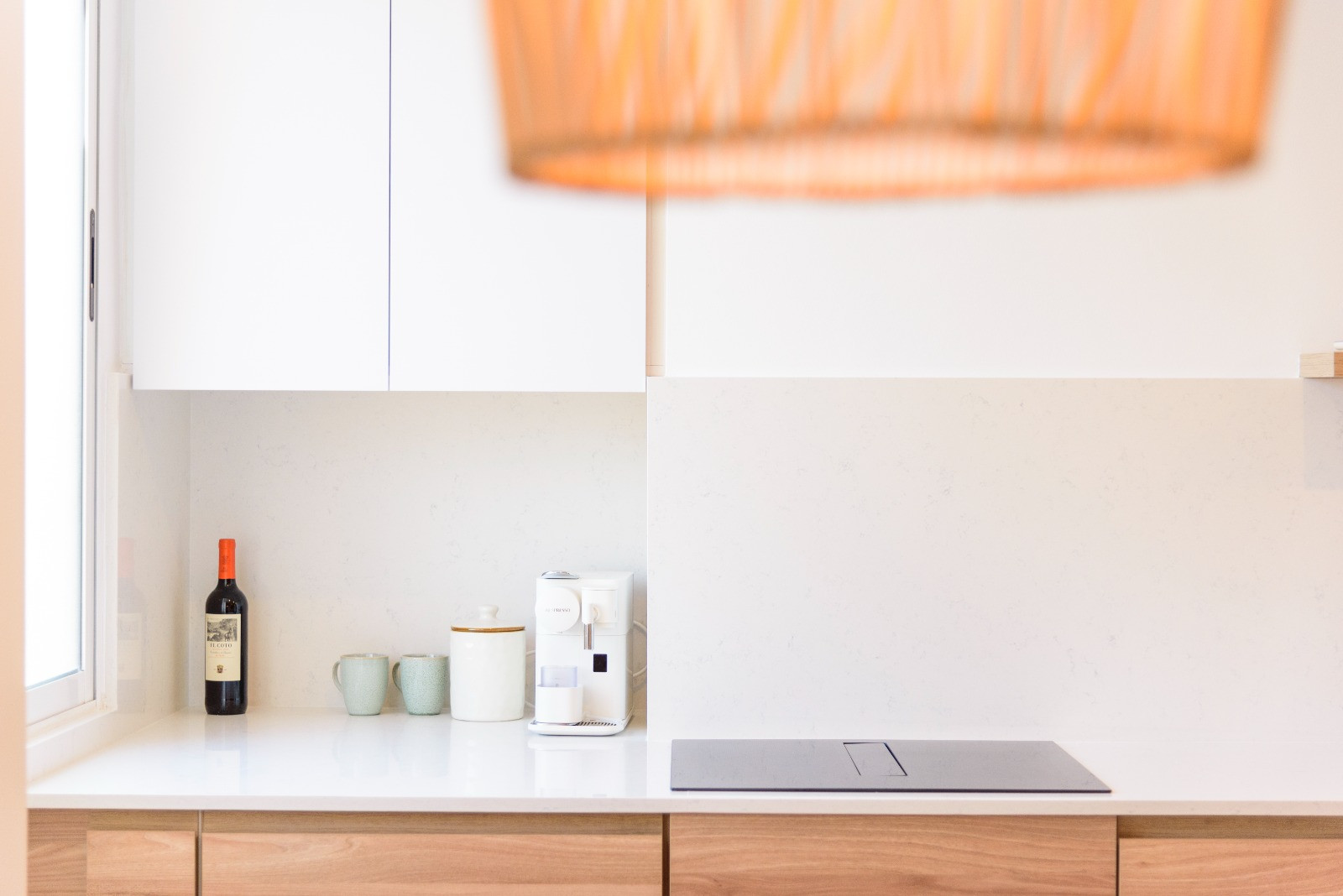 Cuisine contemporaine avec plan de travail en pierre blanche, plaque à induction et meubles en bois. La lumière naturelle illumine l'espace café et les finitions épurées du mobilier.