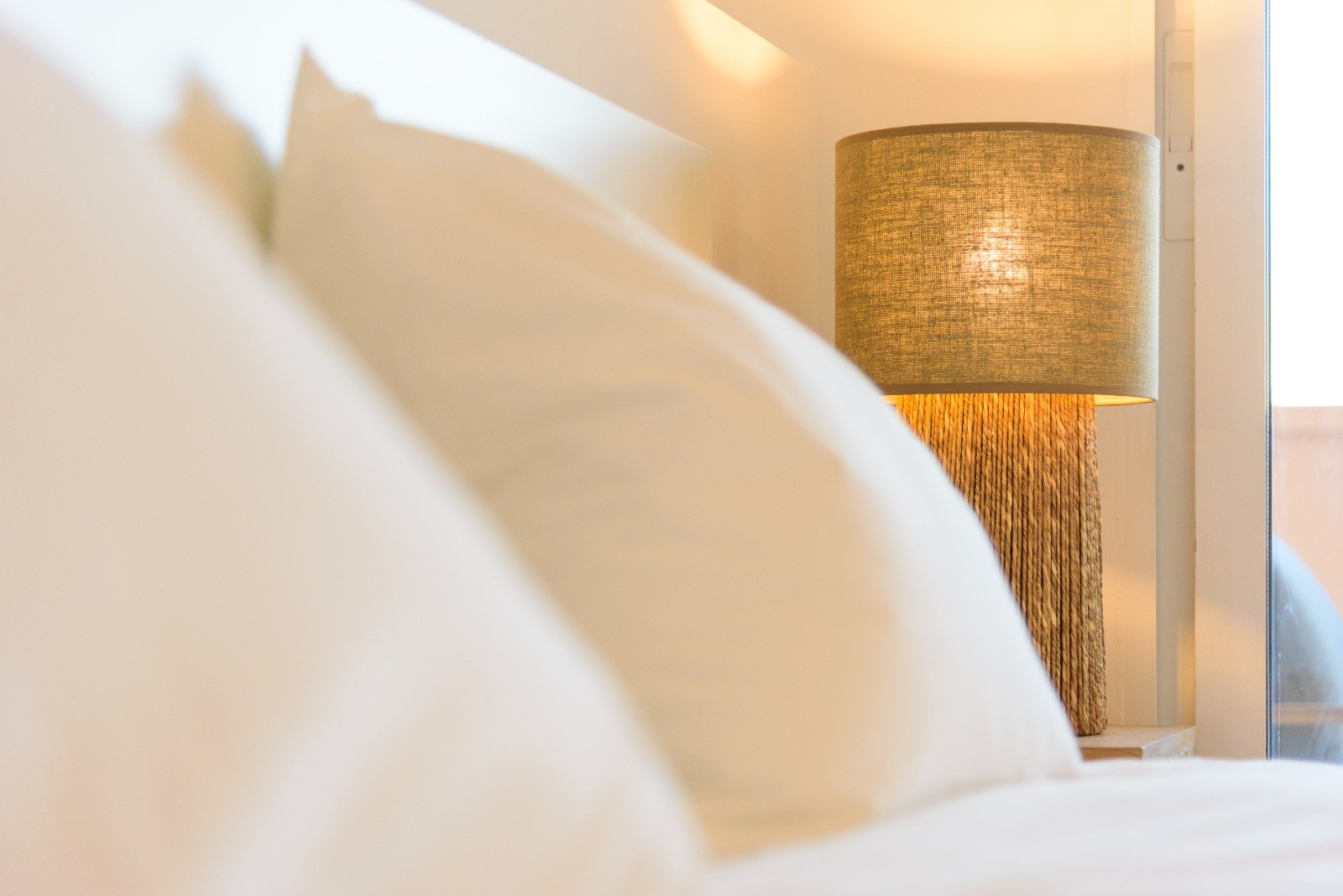 Détail de la chambre avec une lampe de chevet en fibres naturelles et abat-jour en lin, créant une ambiance chaleureuse près du lit aux textiles blancs.