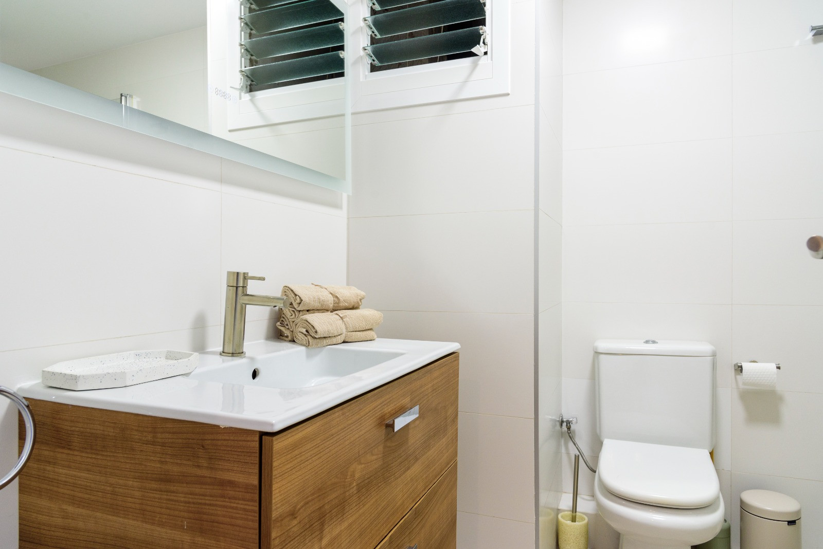 Salle de bain moderne avec meuble sous-vasque en bois, lavabo intégré et carrelage blanc. Comprend des sanitaires contemporains et une fenêtre à persiennes.