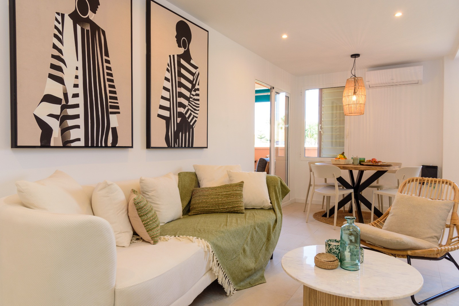 Salon-salle à manger lumineux avec art moderne, table basse en marbre et fauteuil en rotin. Le coin repas dispose d'une suspension en fibres et s'ouvre sur la terrasse ensoleillée.
