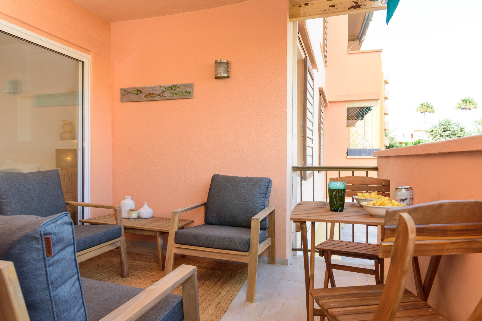Terrasse privée aux murs saumon, équipée d'un salon de jardin en bois avec coussins gris et d'un coin repas. Accès direct à l'intérieur par une grande baie vitrée.