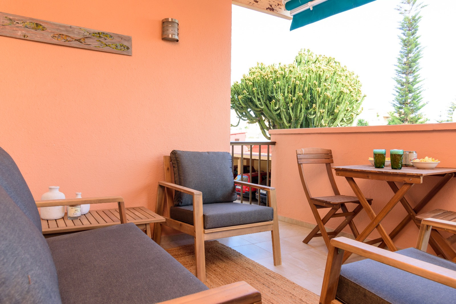 Terrasse privée avec mobilier en bois, coussins gris et coin repas extérieur, offrant une vue sur la végétation environnante sous un store banne bleu.