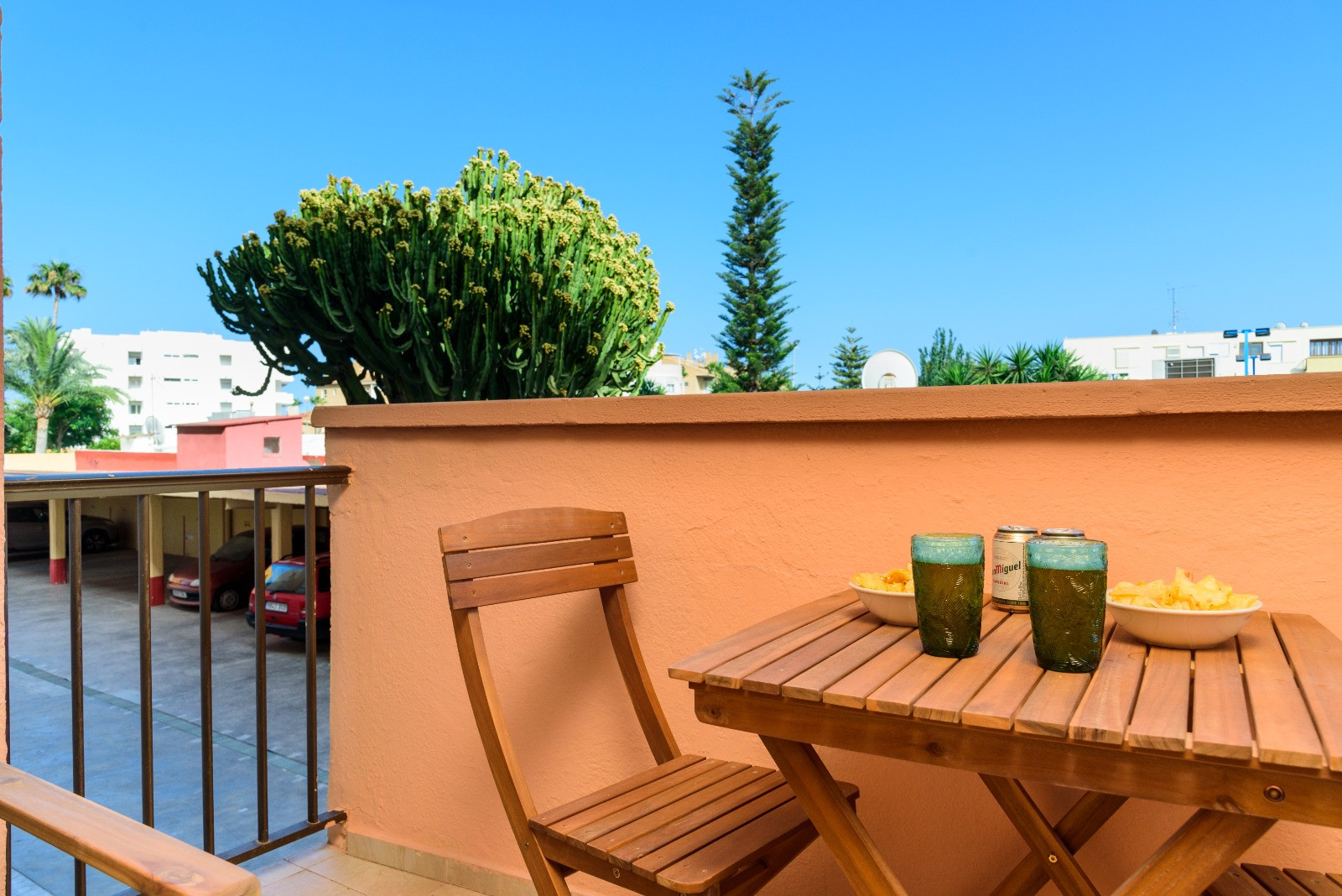 Terrasse privée avec mobilier en bois, idéale pour des apéritifs en plein air. Elle offre une vue sur des jardins avec cactus et palmiers sous un ciel bleu azur.