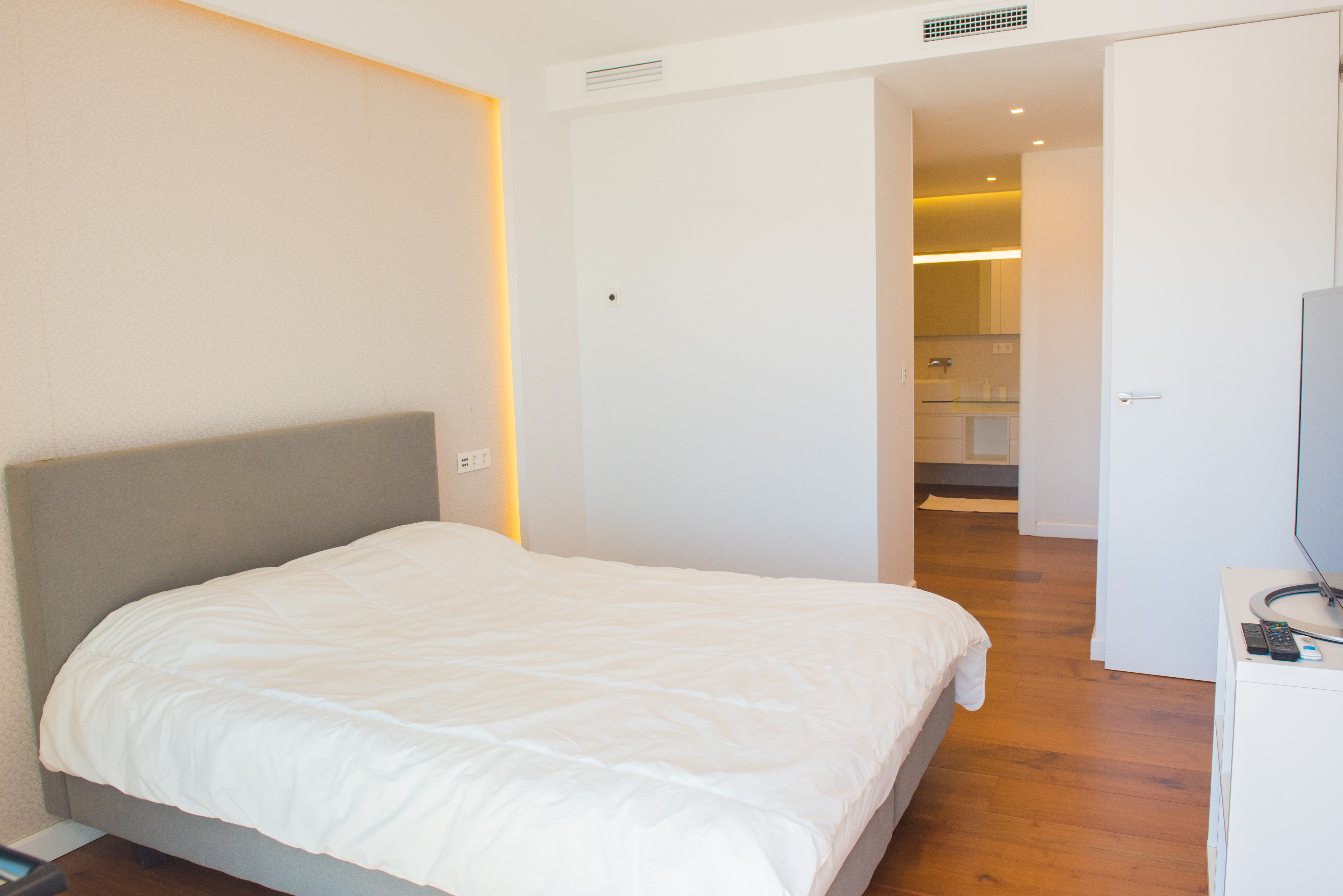 Chambre principale avec tête de lit grise, éclairage LED indirect au mur, parquet chaleureux et vue sur la salle de bain attenante au design épuré.