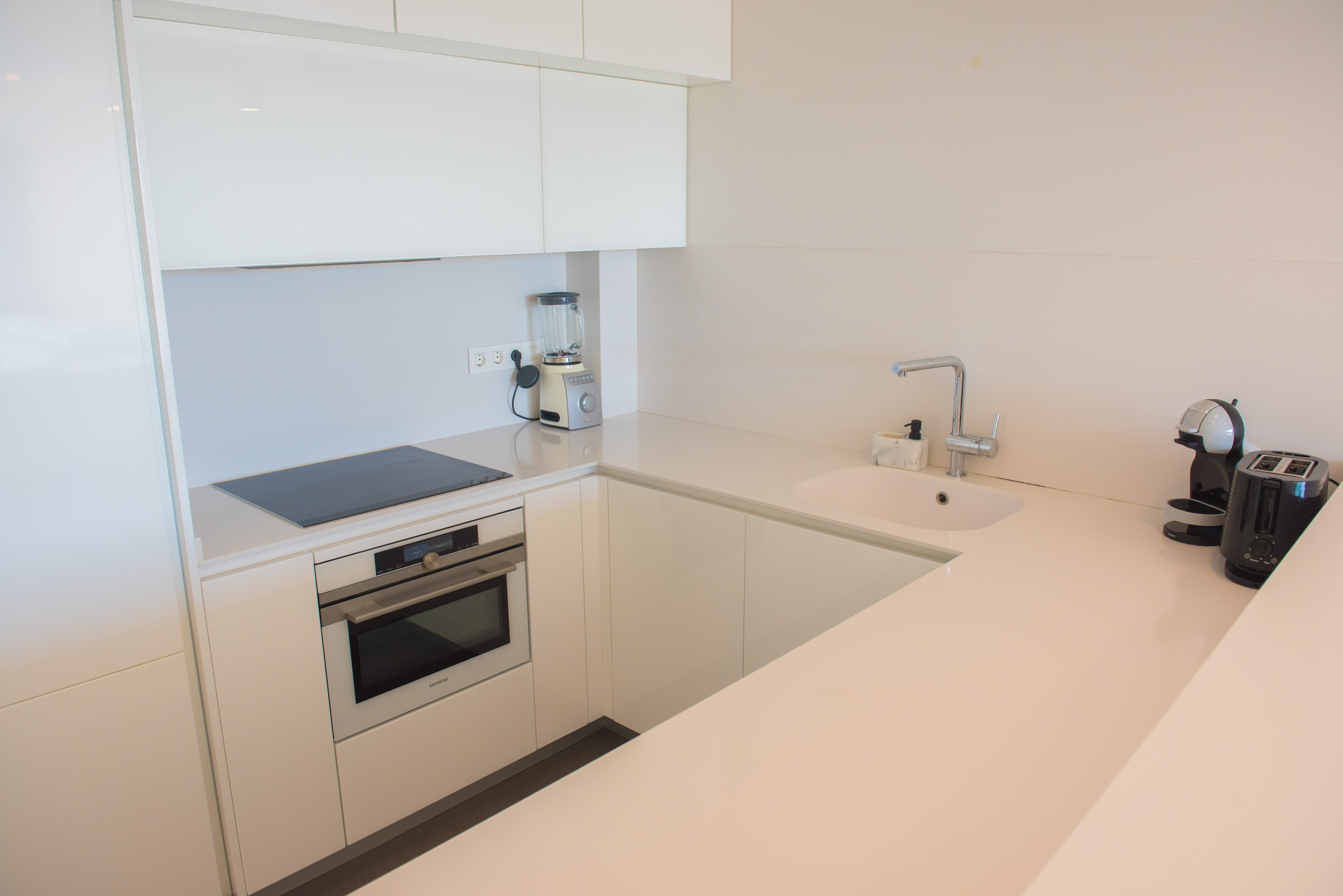 Cuisine minimaliste blanc brillant avec plan de travail en quartz, four Siemens, plaque à induction et petit électroménager haut de gamme intégré.