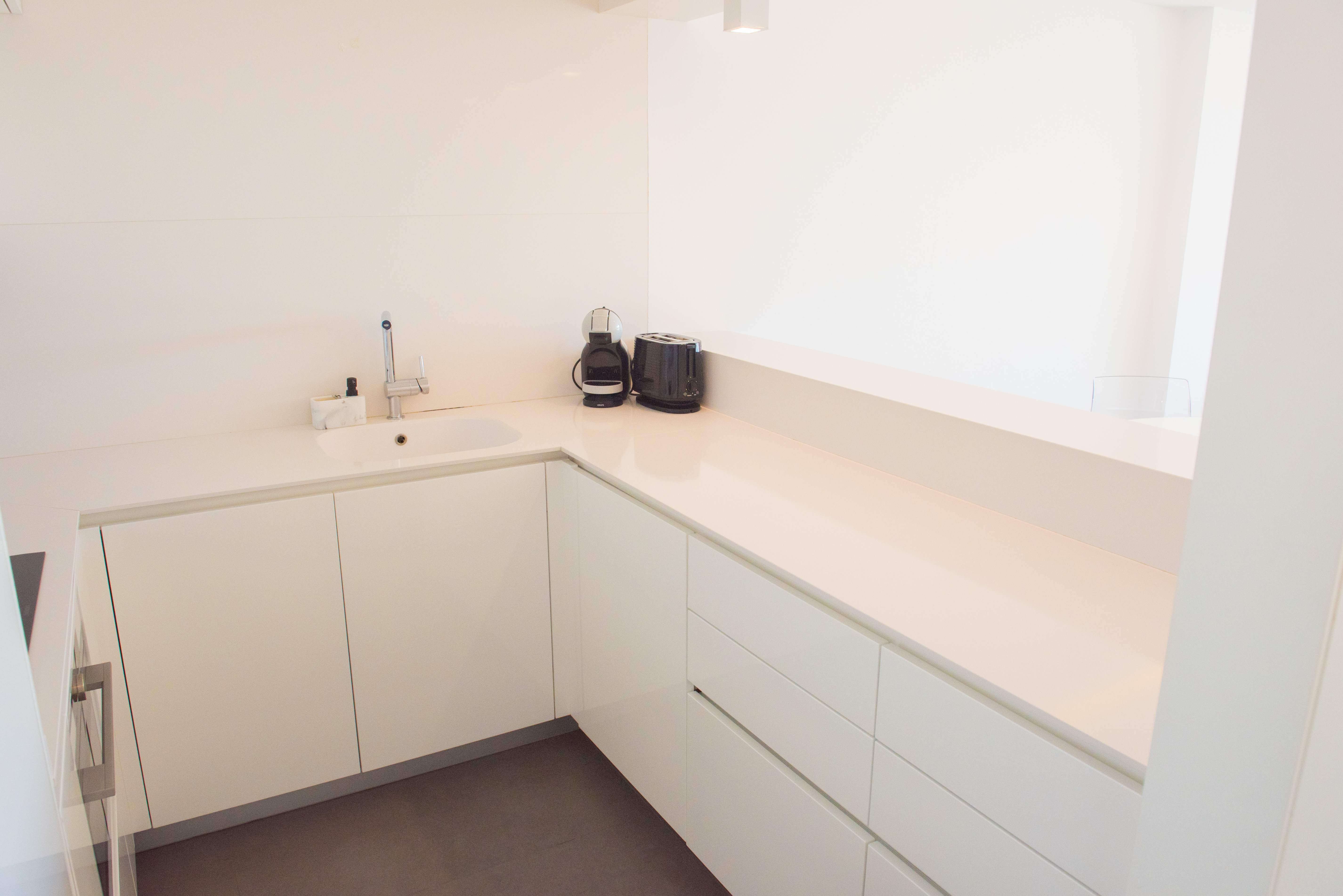 Cuisine minimaliste moderne en blanc brillant avec plan de travail en quartz, évier intégré et carrelage sombre contrasté.