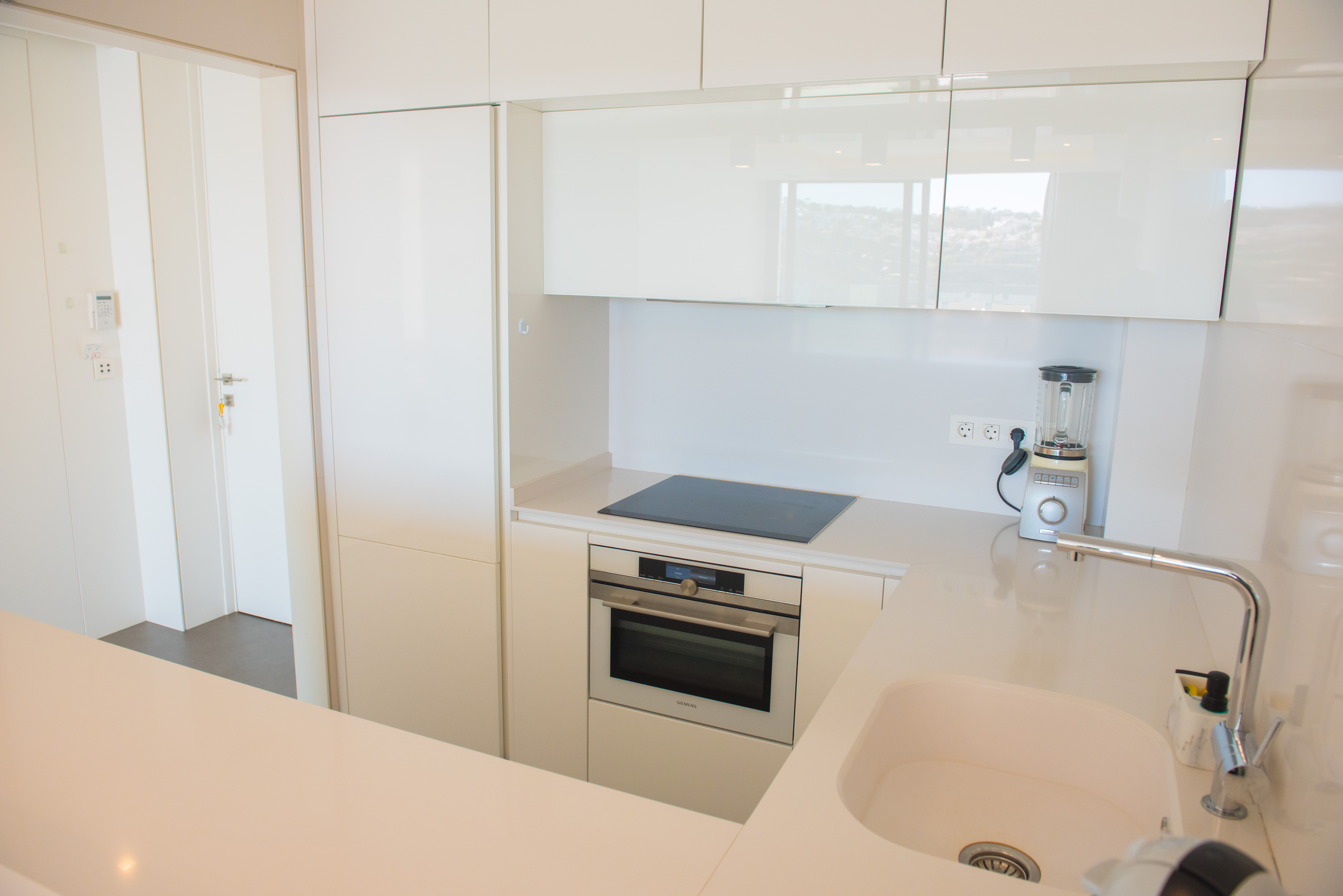 Cuisine moderne avec placards blancs brillants, four Siemens encastré, plaque à induction et plan de travail en quartz blanc avec évier sous plan.