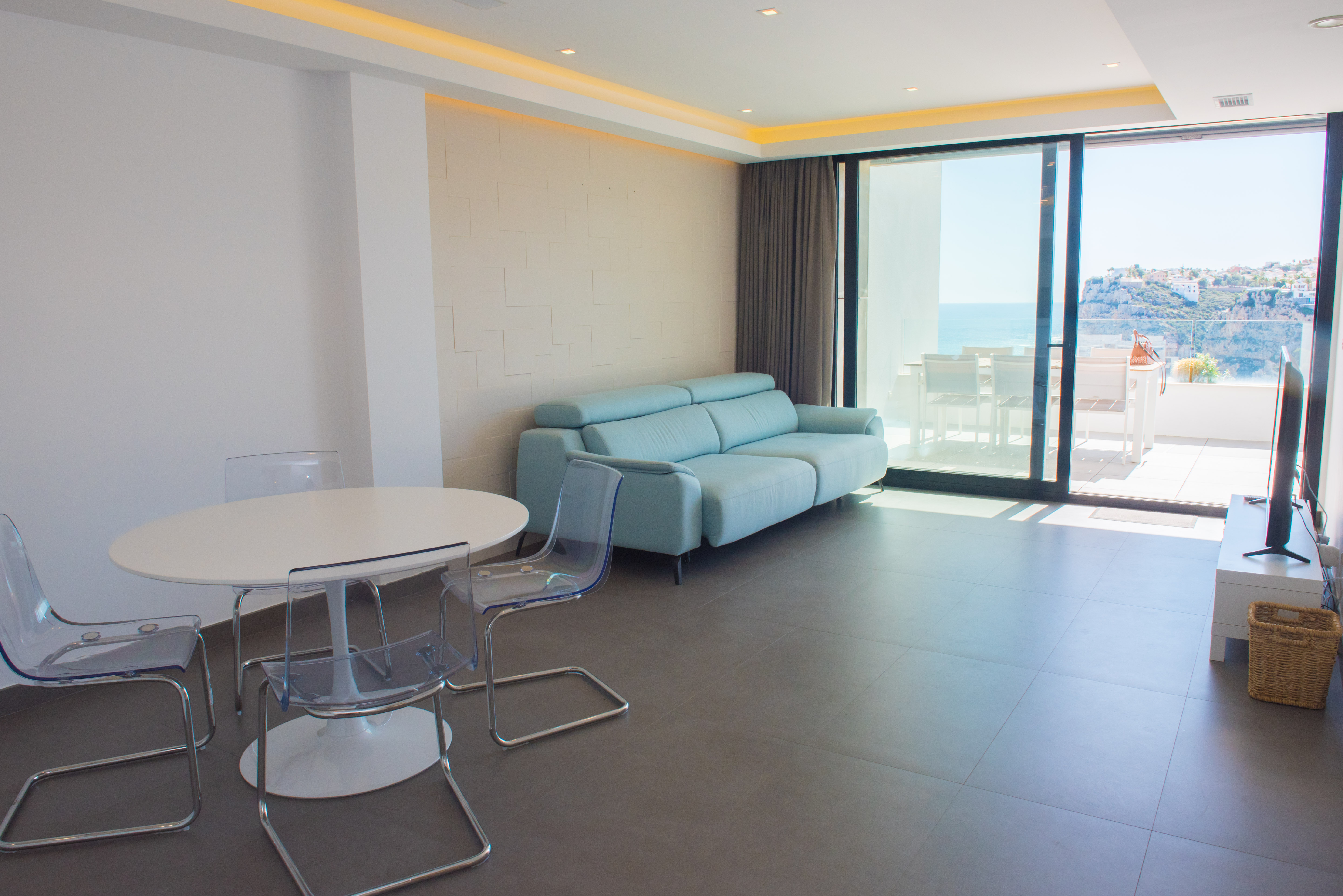 Espace de vie avec canapé bleu pastel, chaises en acrylique et éclairage indirect. Les grandes baies vitrées offrent une transition fluide vers la terrasse vue mer.