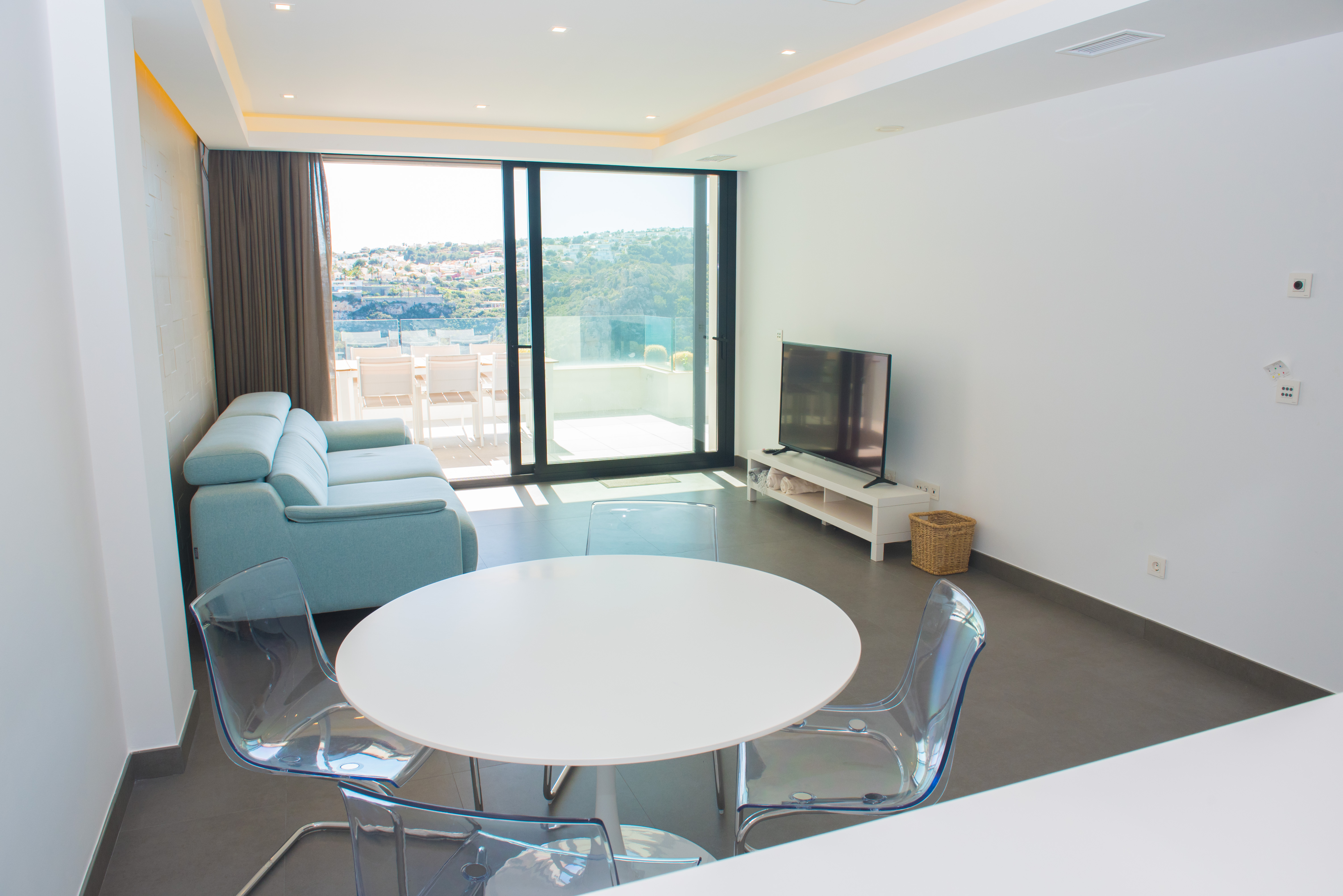Espace de vie minimaliste avec table circulaire blanche, chaises en acrylique et canapé bleu clair, s'ouvrant sur une terrasse avec vue panoramique par de grandes baies.