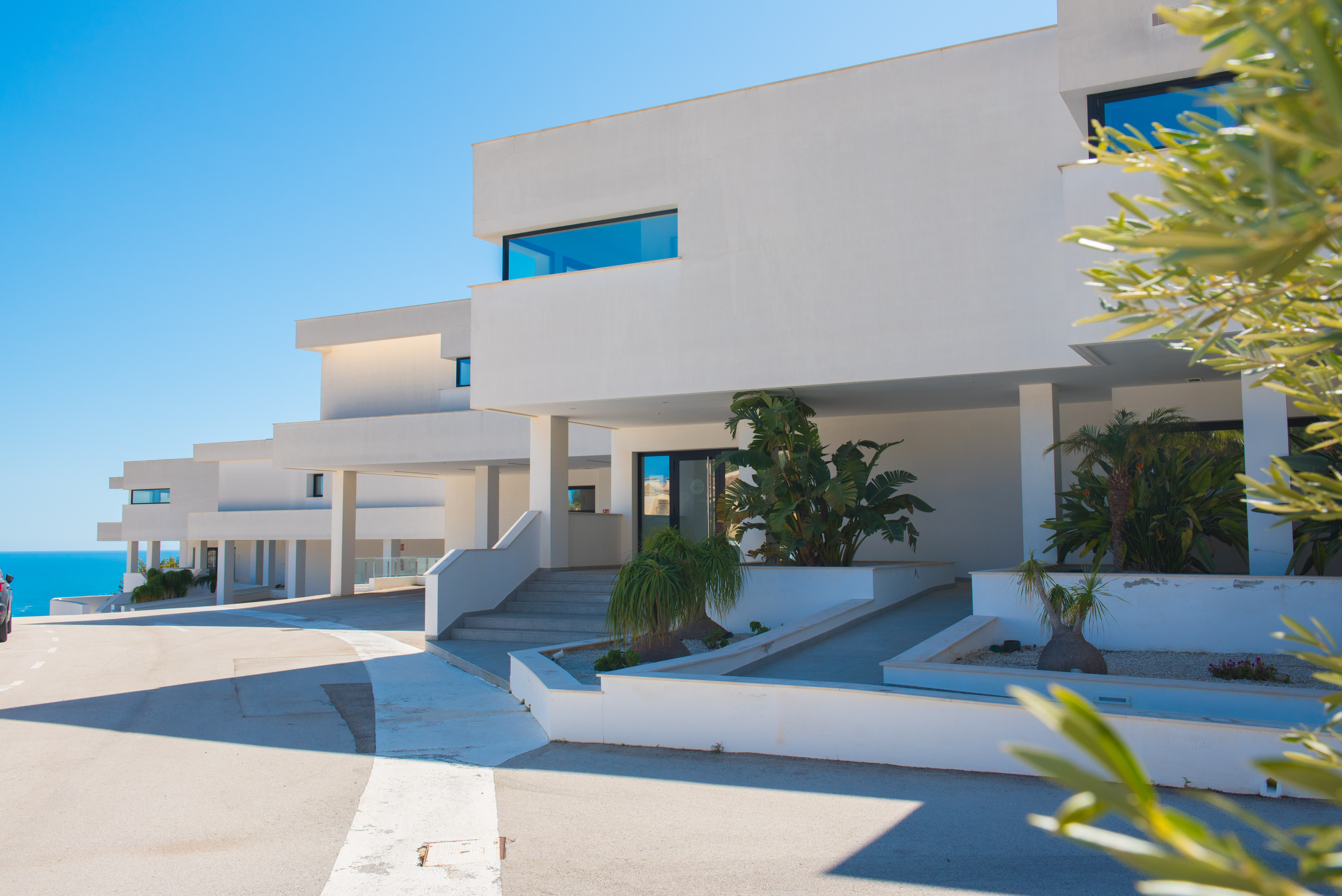 Façade minimaliste d'un blanc pur avec accès par escaliers et rampes, jardinières intégrées de plantes tropicales et lignes architecturales épurées.