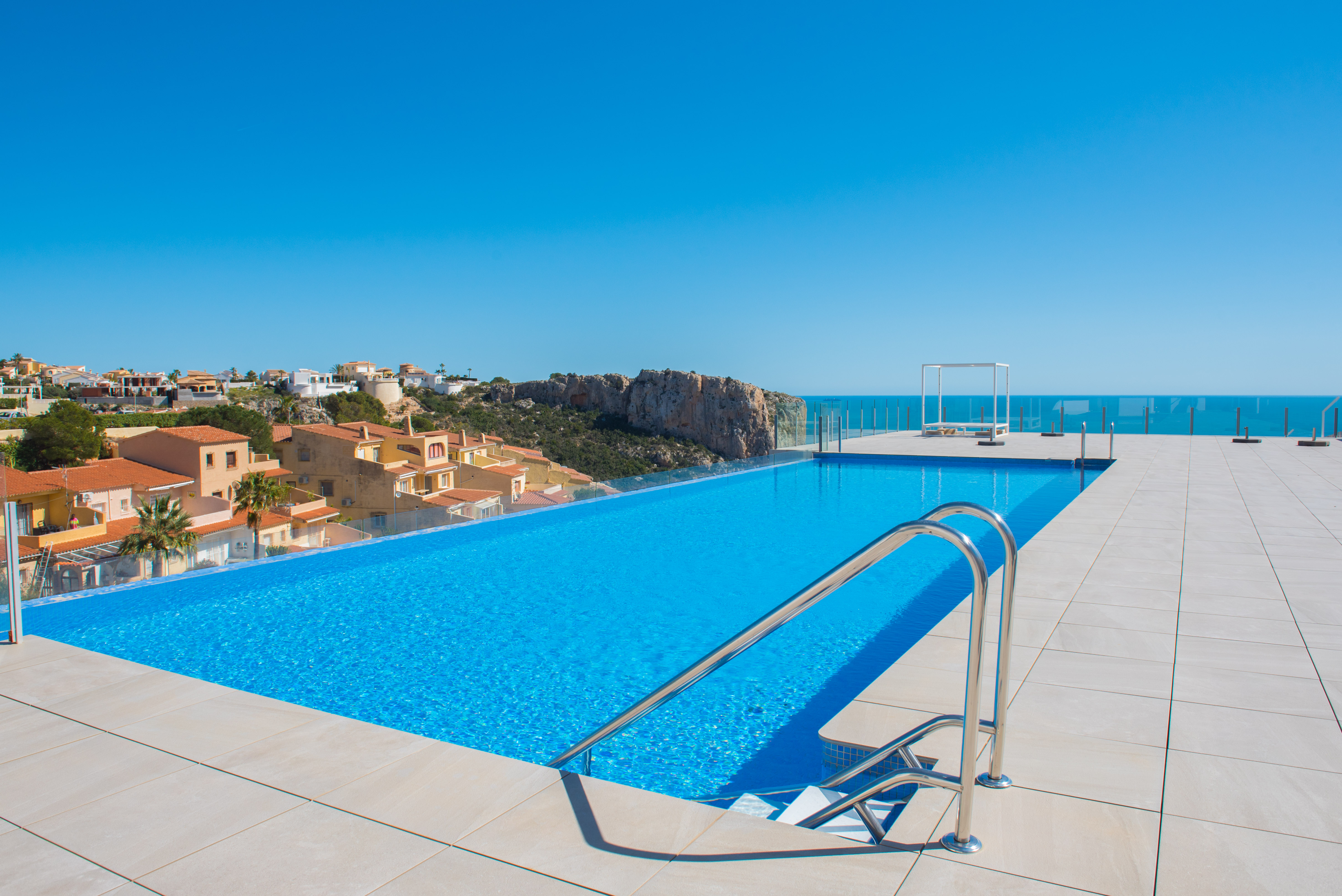 Piscine à débordement avec garde-corps en verre et vue panoramique sur la mer et les falaises. La terrasse spacieuse est revêtue de dalles claires et dispose d'un lit de repos moderne.