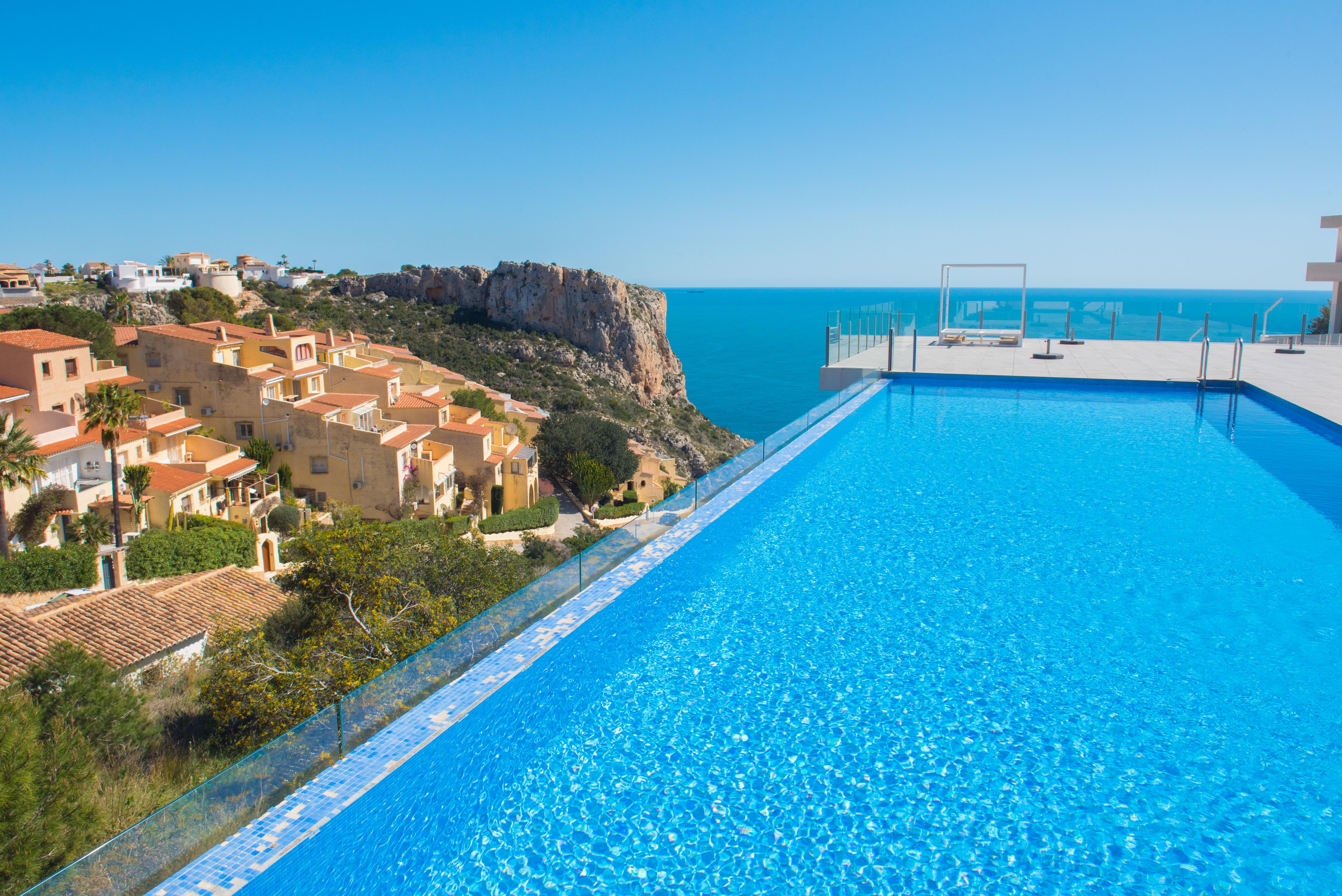 Piscine à débordement avec mosaïques bleues et garde-corps vitrés, offrant une vue sur la mer Méditerranée, les falaises et les villas environnantes.