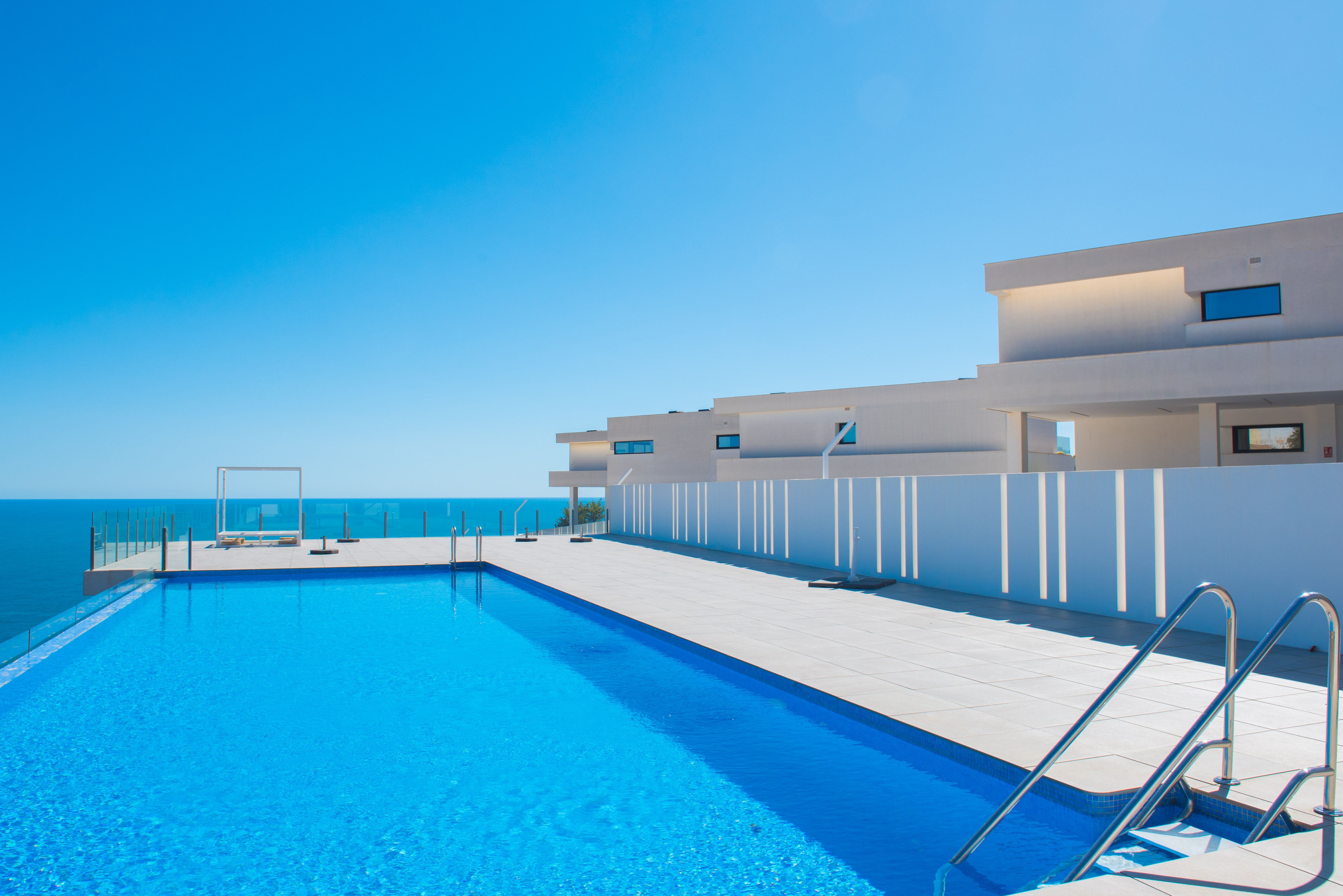 Piscine à débordement face à la mer, avec une vaste terrasse carrelée, des garde-corps en verre et des bâtiments blancs au design contemporain.