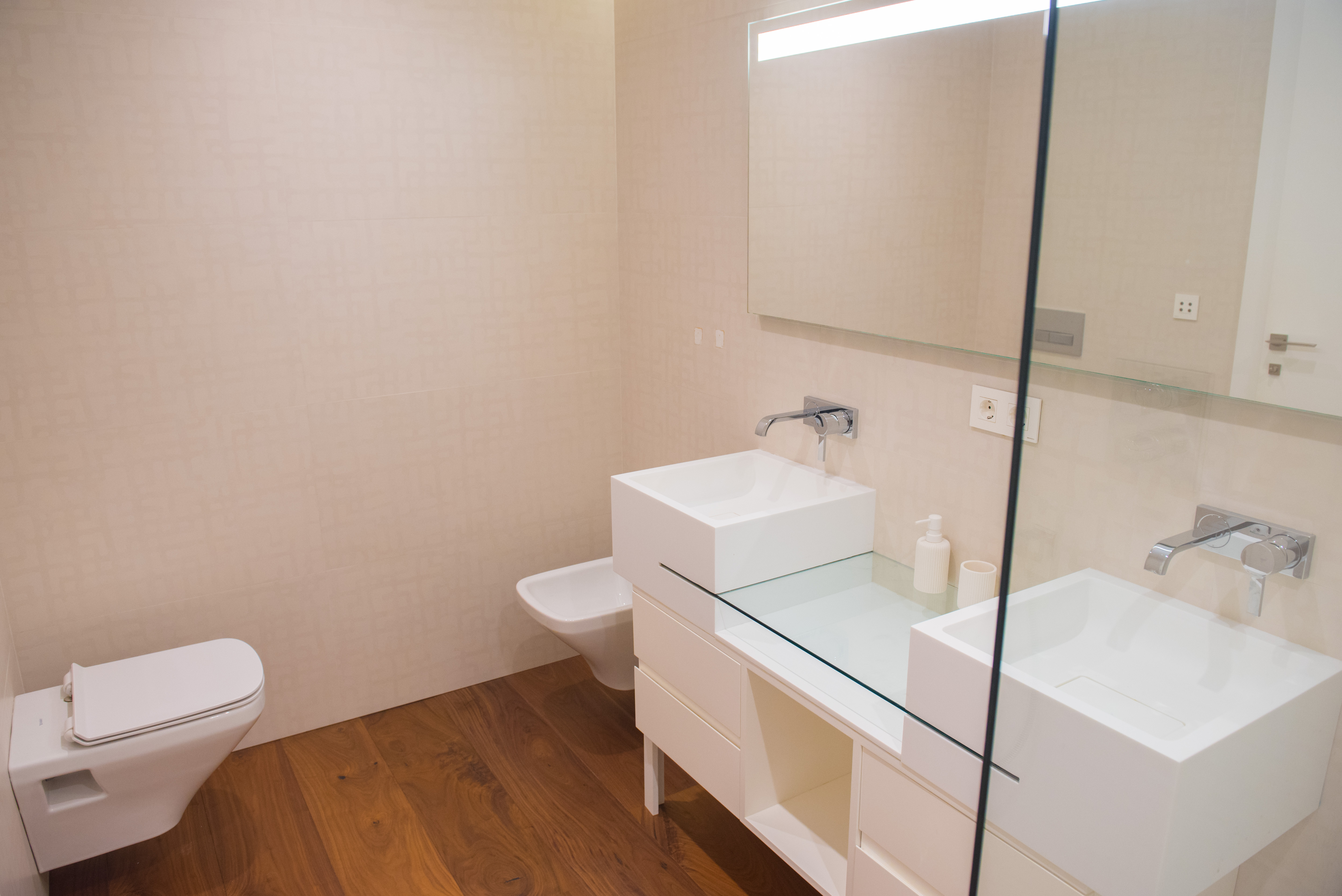 Salle de bain contemporaine avec double vasque carrée, plan de travail en verre et robinetterie murale. Équipée de sanitaires suspendus et d'un sol en bois.