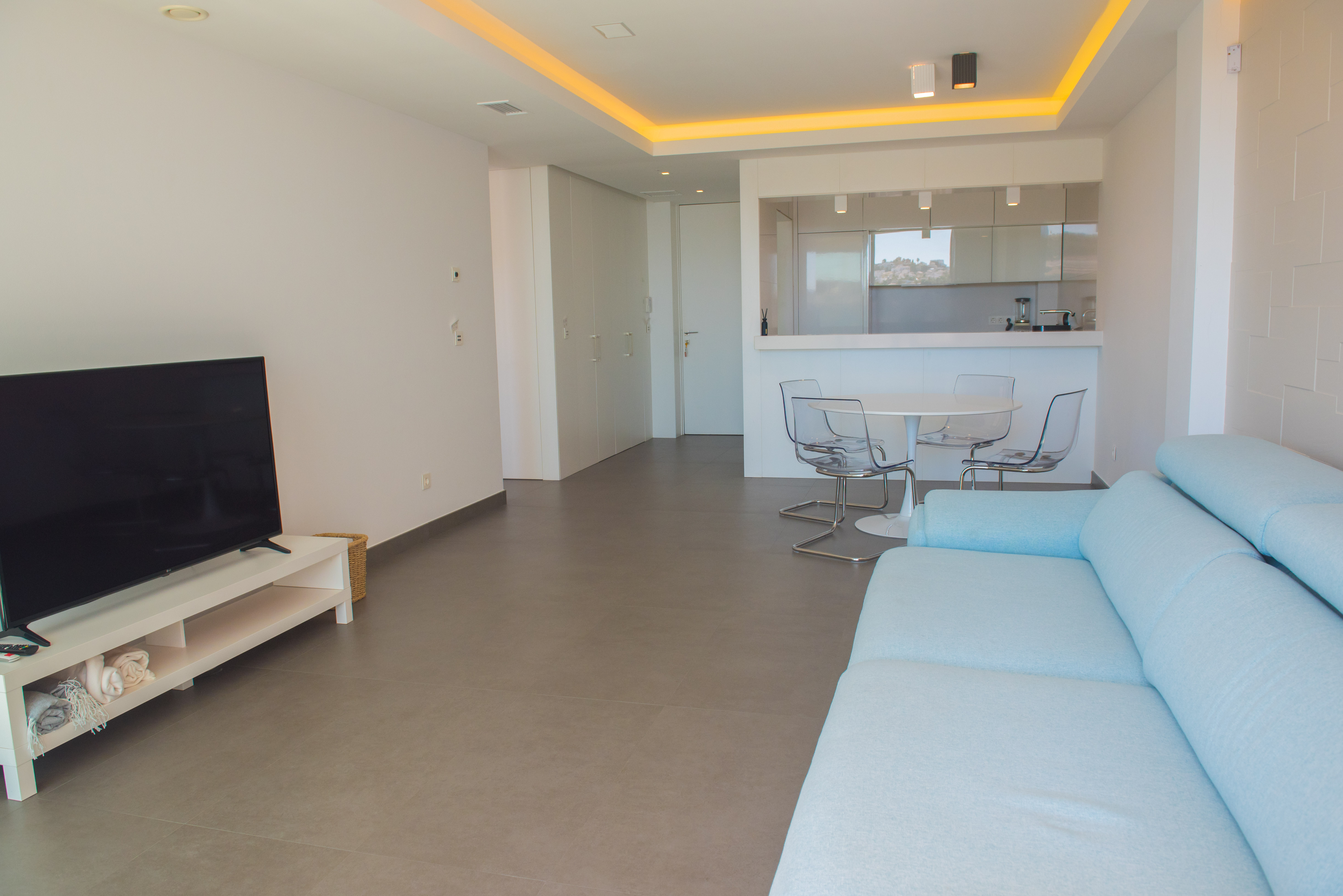 Salon à aire ouverte avec canapé bleu clair, salle à manger aux chaises transparentes et cuisine intégrée sous un plafond à éclairage LED.