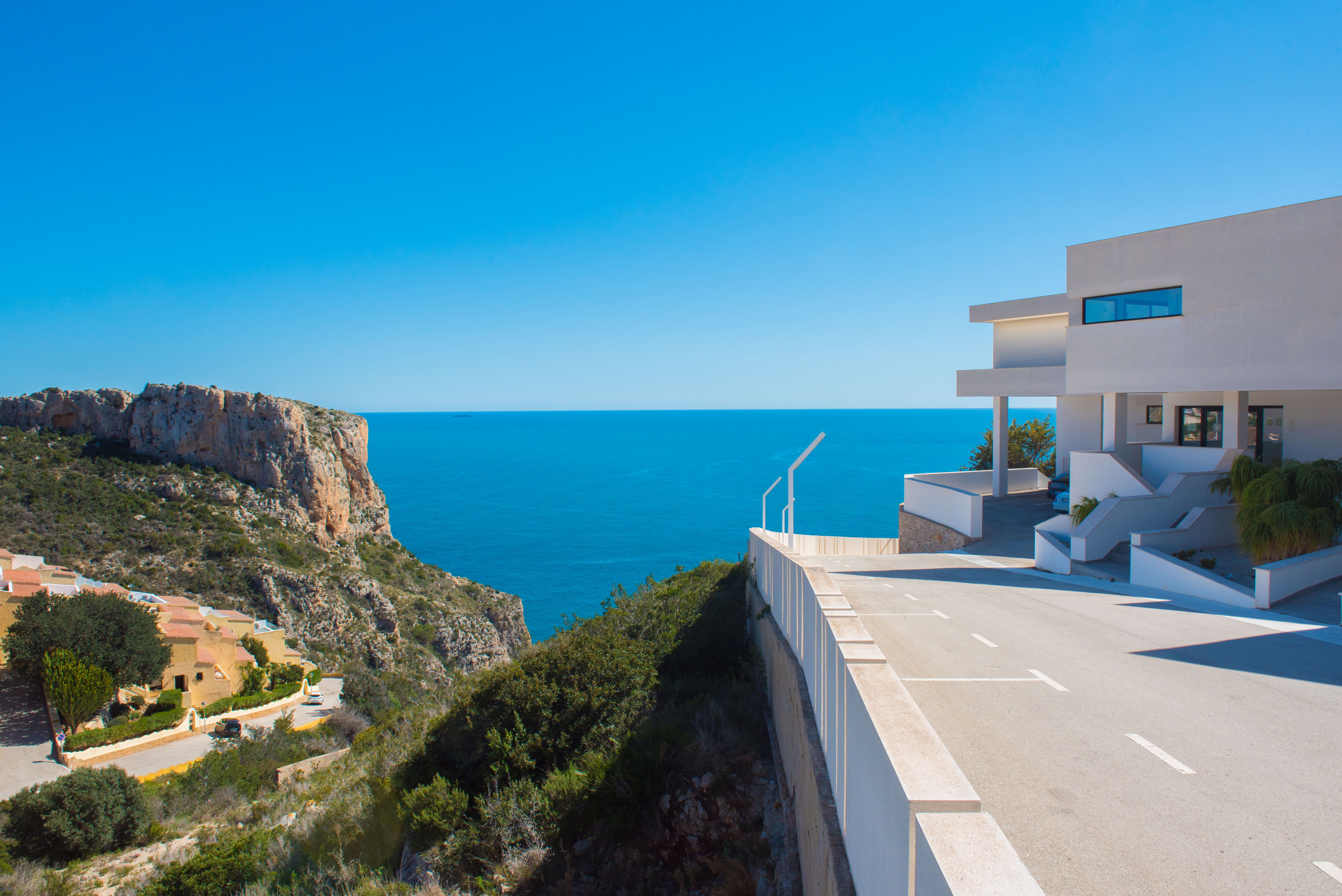 Vue extérieure du complexe à l'architecture minimaliste blanche, situé sur une falaise avec accès pavé et vue panoramique sur l'horizon méditerranéen.