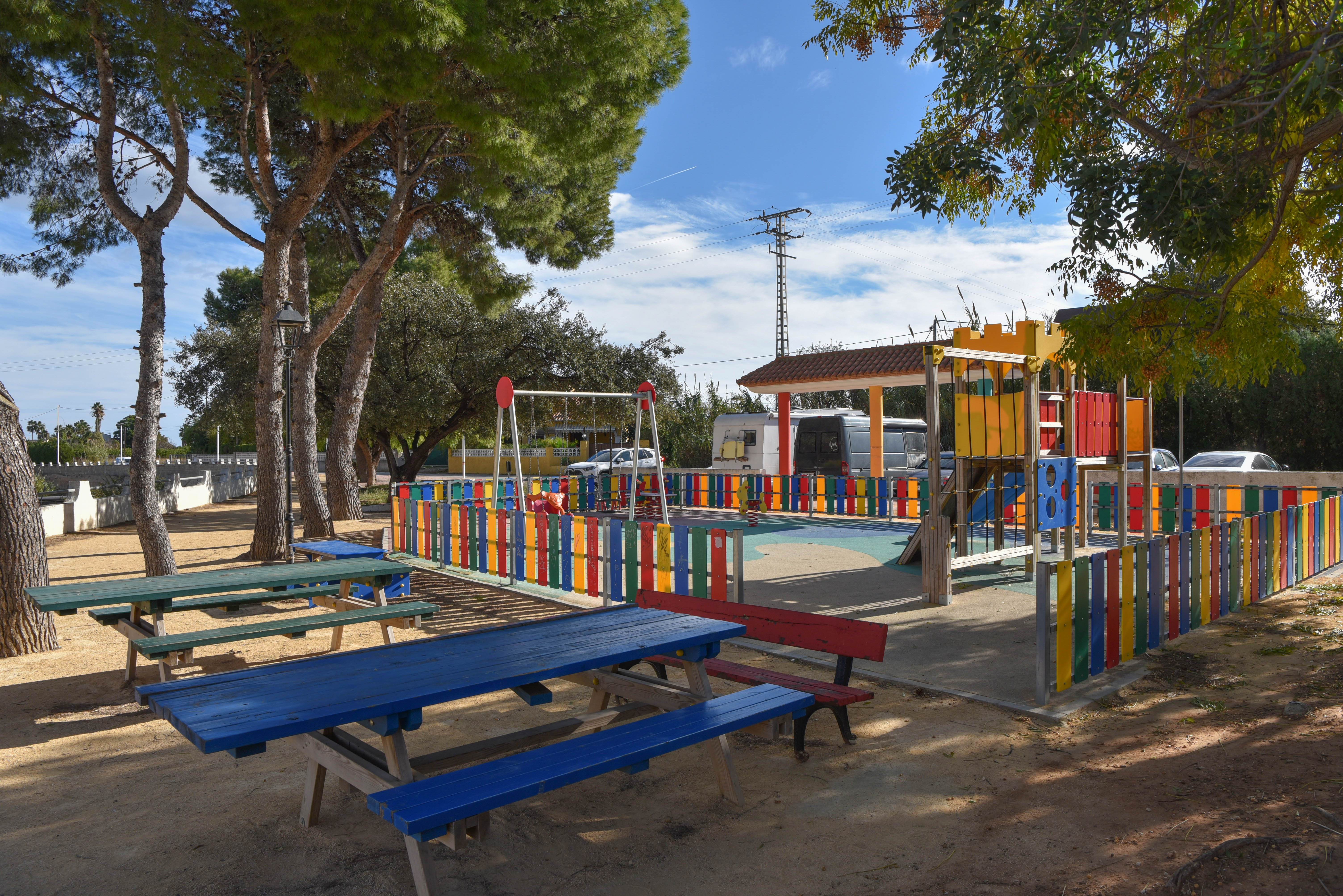 Aire de jeux clôturée pour enfants avec structures colorées et tables de pique-nique en bois, ombragée par de grands pins sous un ciel dégagé.