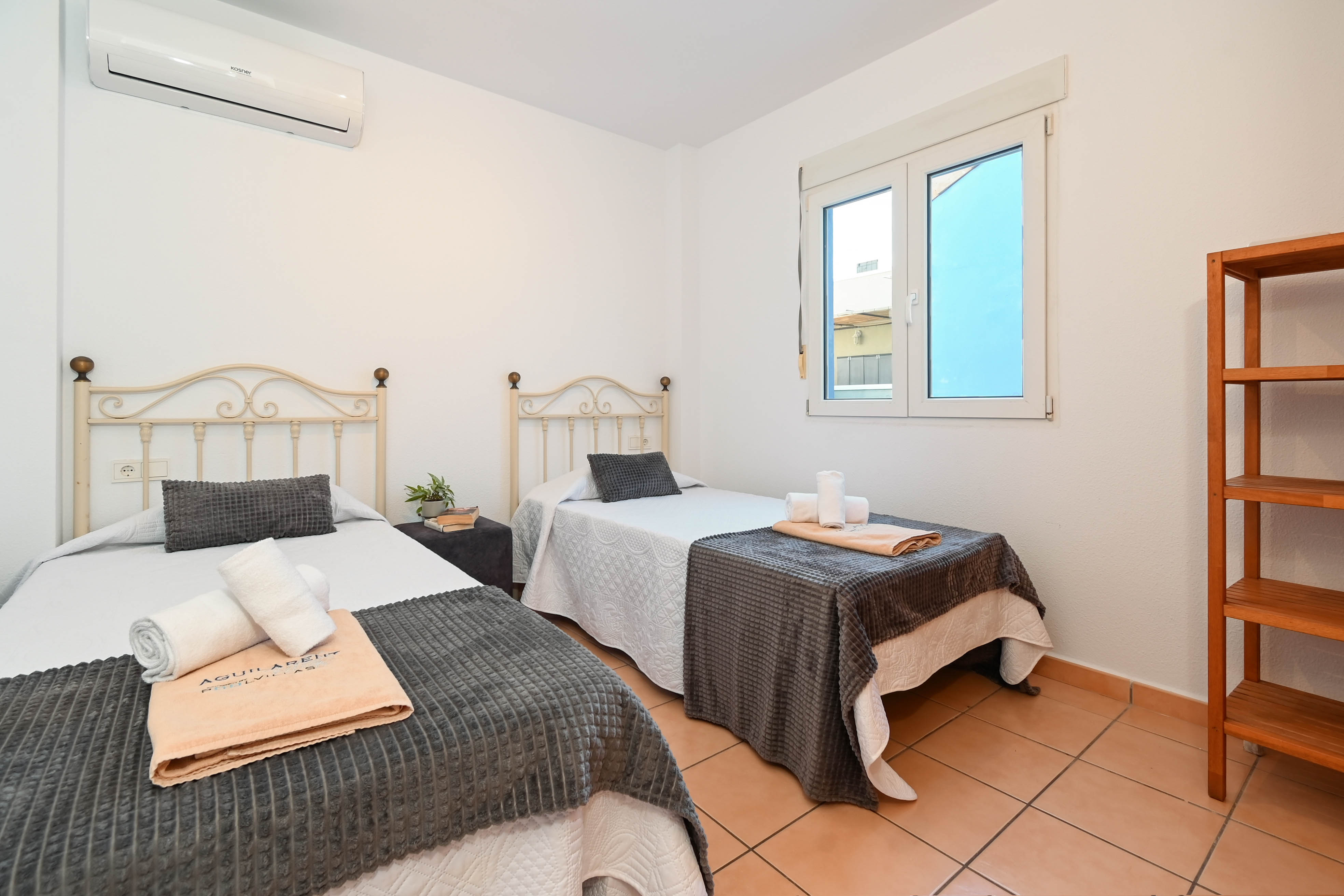 Chambre avec deux lits simples, têtes de lit en fer forgé, sol en terre cuite et climatisation. Équipée d'une étagère en bois et d'une fenêtre à cadre blanc.