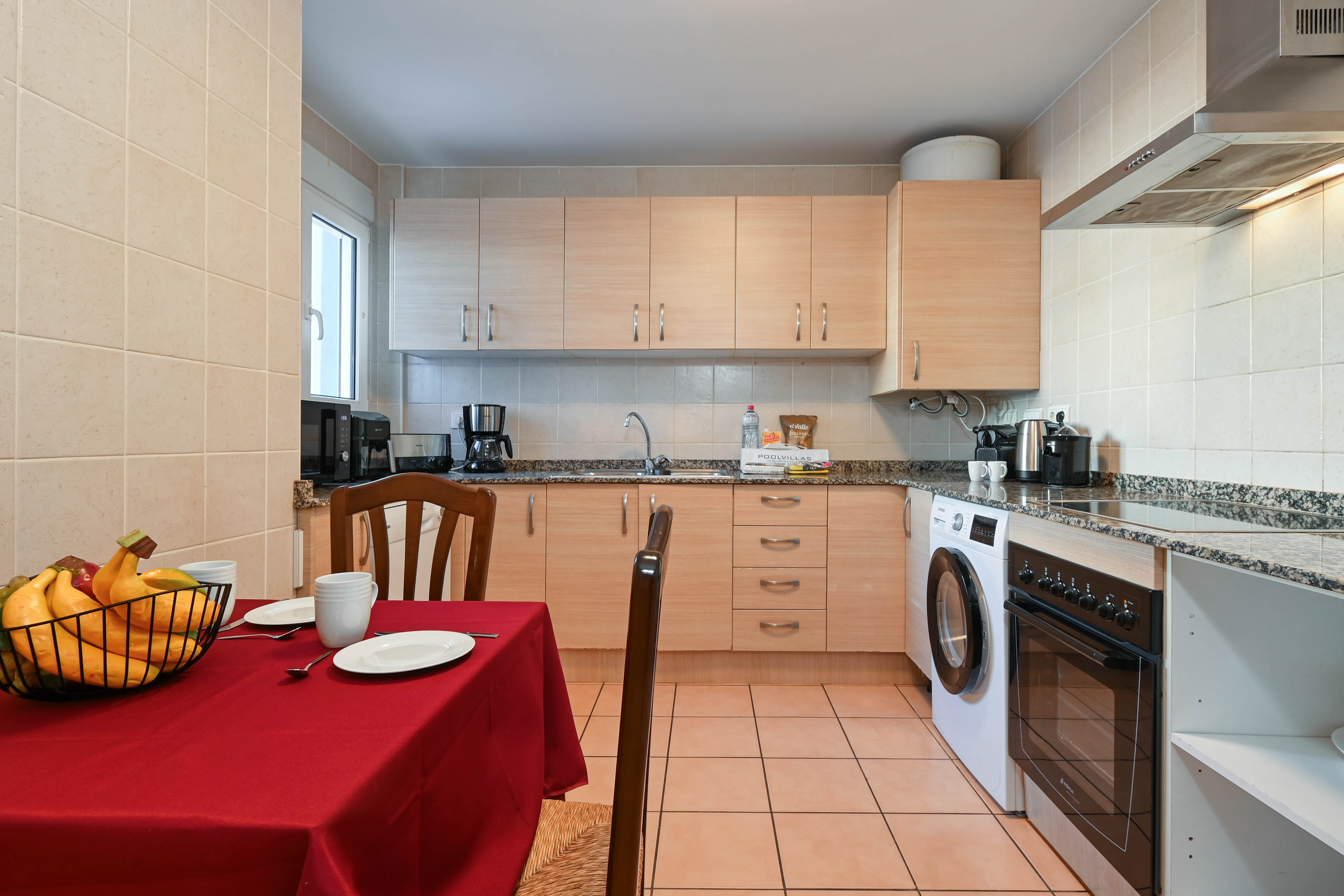 Cuisine équipée avec placards en bois clair, plans de travail en granit et appareils intégrés, incluant un coin repas avec nappe rouge.
