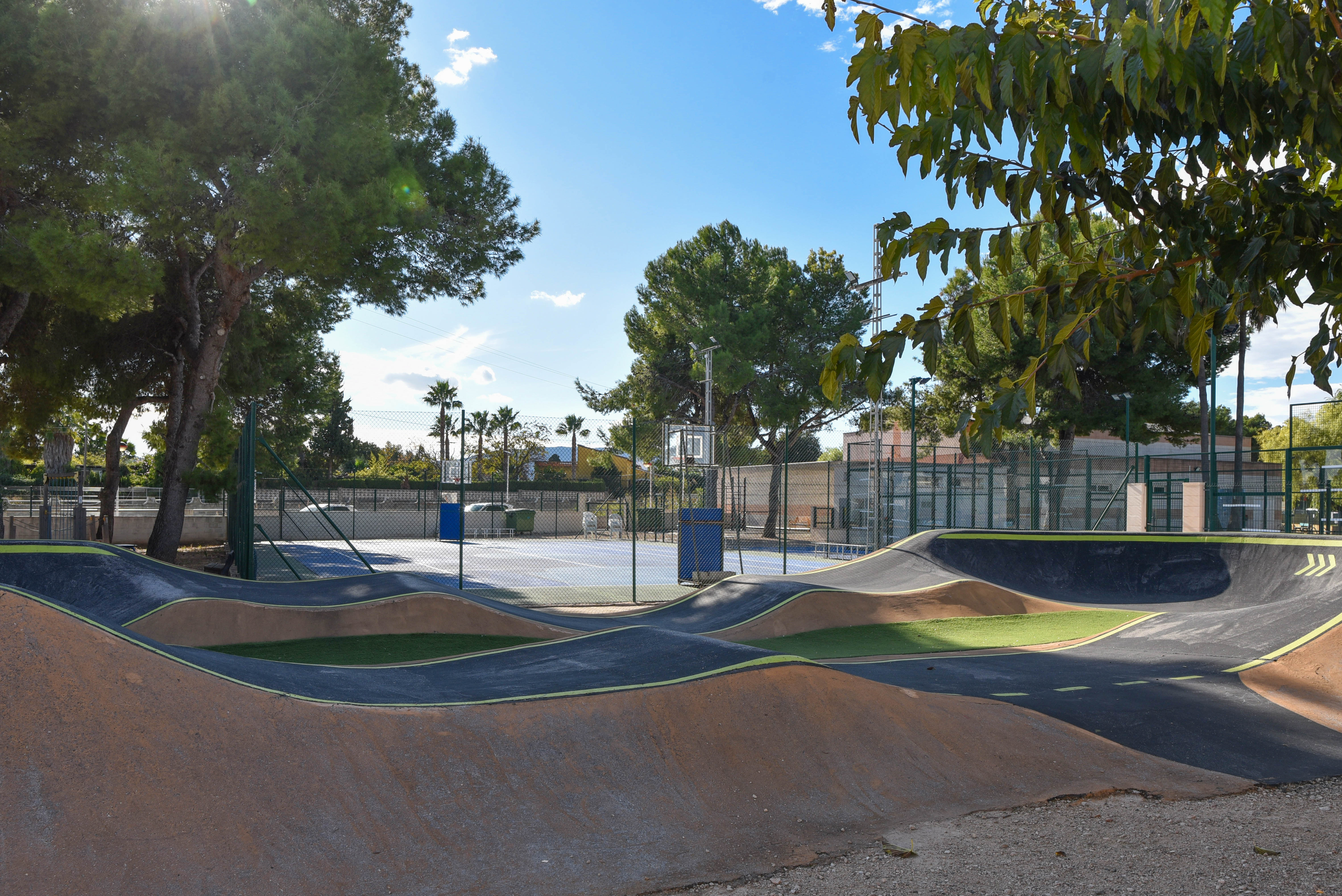 Piste de pump track en asphalte avec rampes ondulées située à côté d'un terrain multisports clôturé, entourée de pins dans un espace de loisirs.