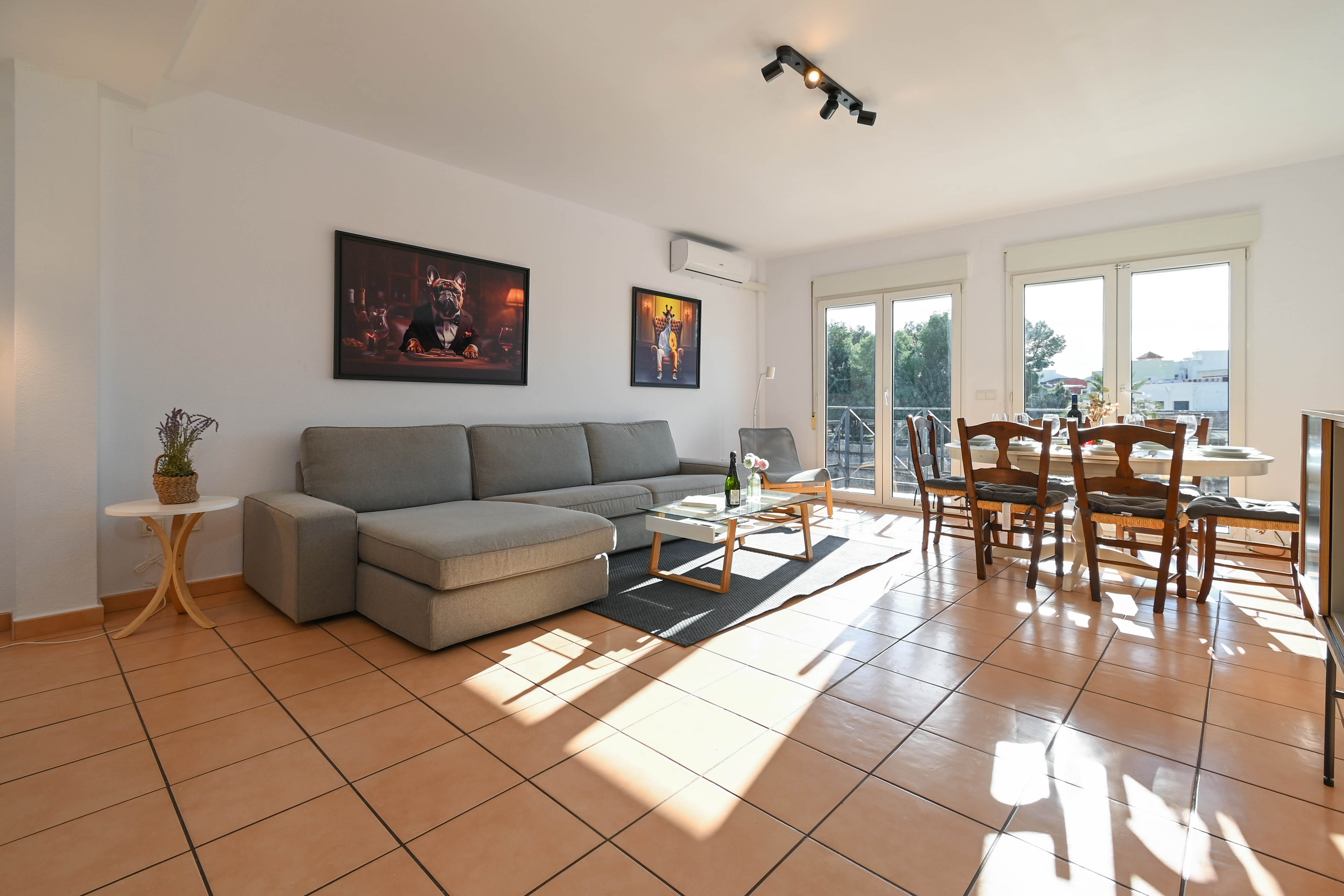 Salon-salle à manger lumineux avec carrelage en terre cuite, canapé d'angle gris et grandes baies vitrées ouvrant sur l'extérieur.