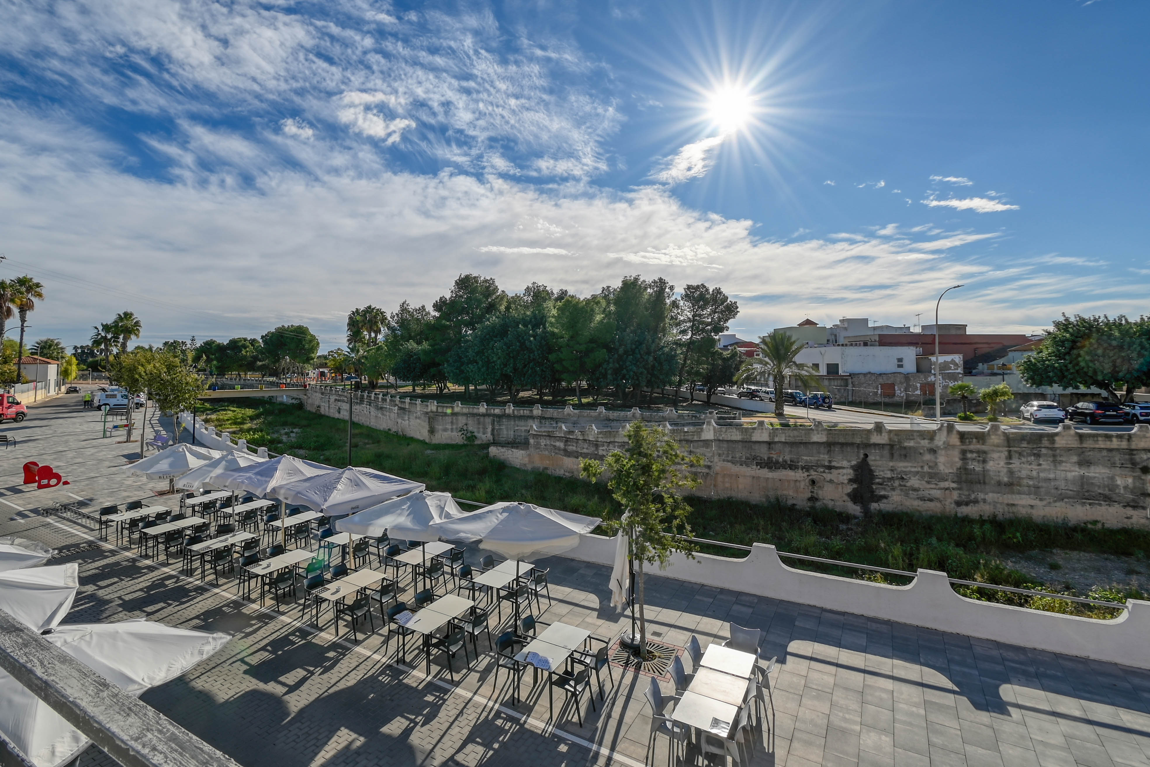 Terrasse extérieure avec parasols blancs et mobilier moderne, offrant une vue sur un canal bordé de murs en pierre et une zone arborée ensoleillée.
