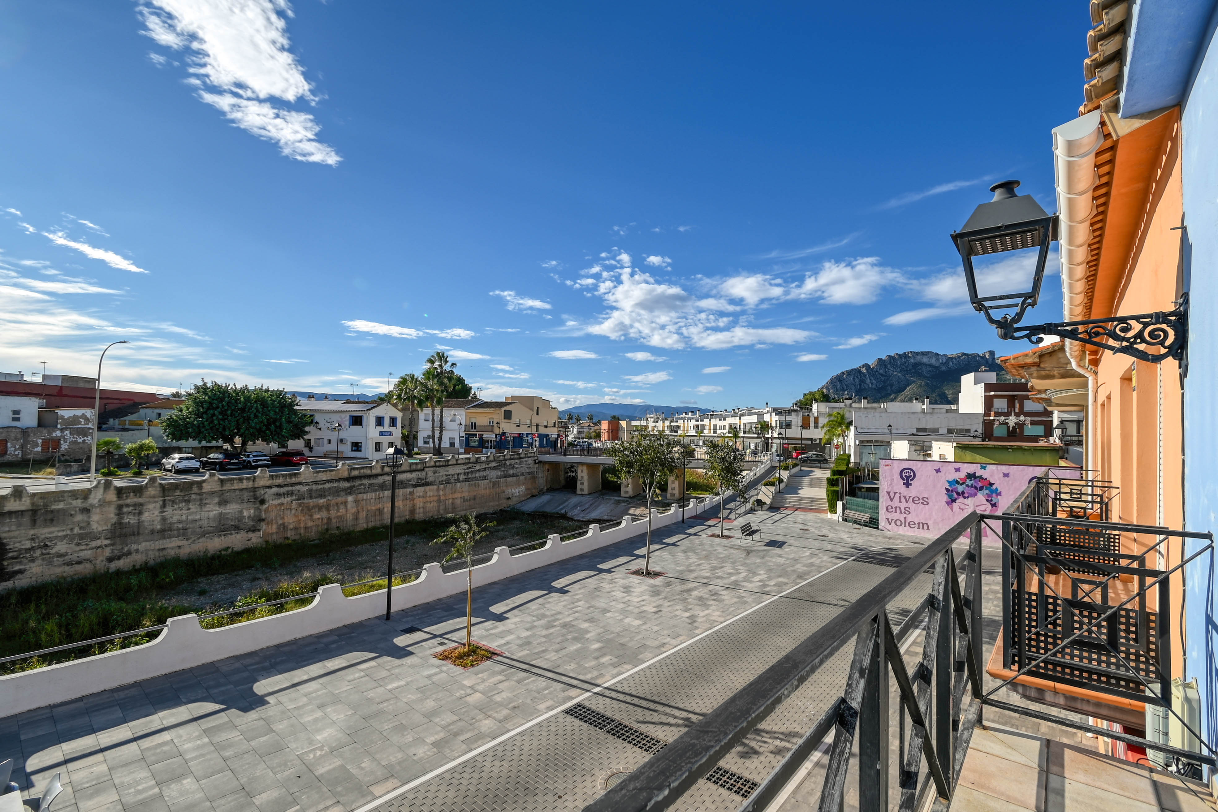 Vue depuis le balcon avec balustrade en fer, donnant sur une place piétonne pavée, le lit de la rivière et les montagnes de la Marina Alta.