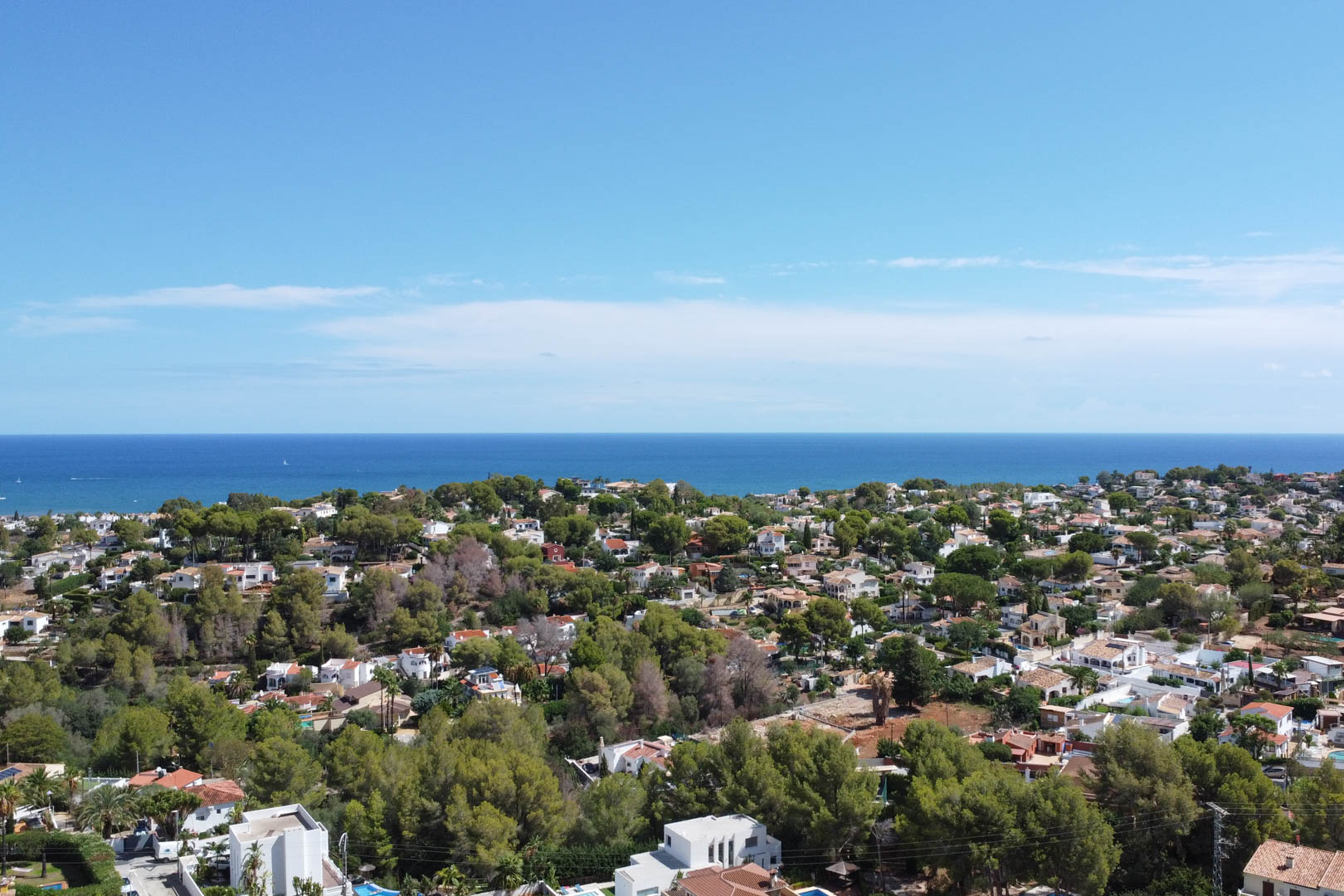 Vue aérienne panoramique d'une zone résidentielle à Dénia, présentant des villas blanches entourées de pins méditerranéens avec l'horizon marin en arrière-plan.