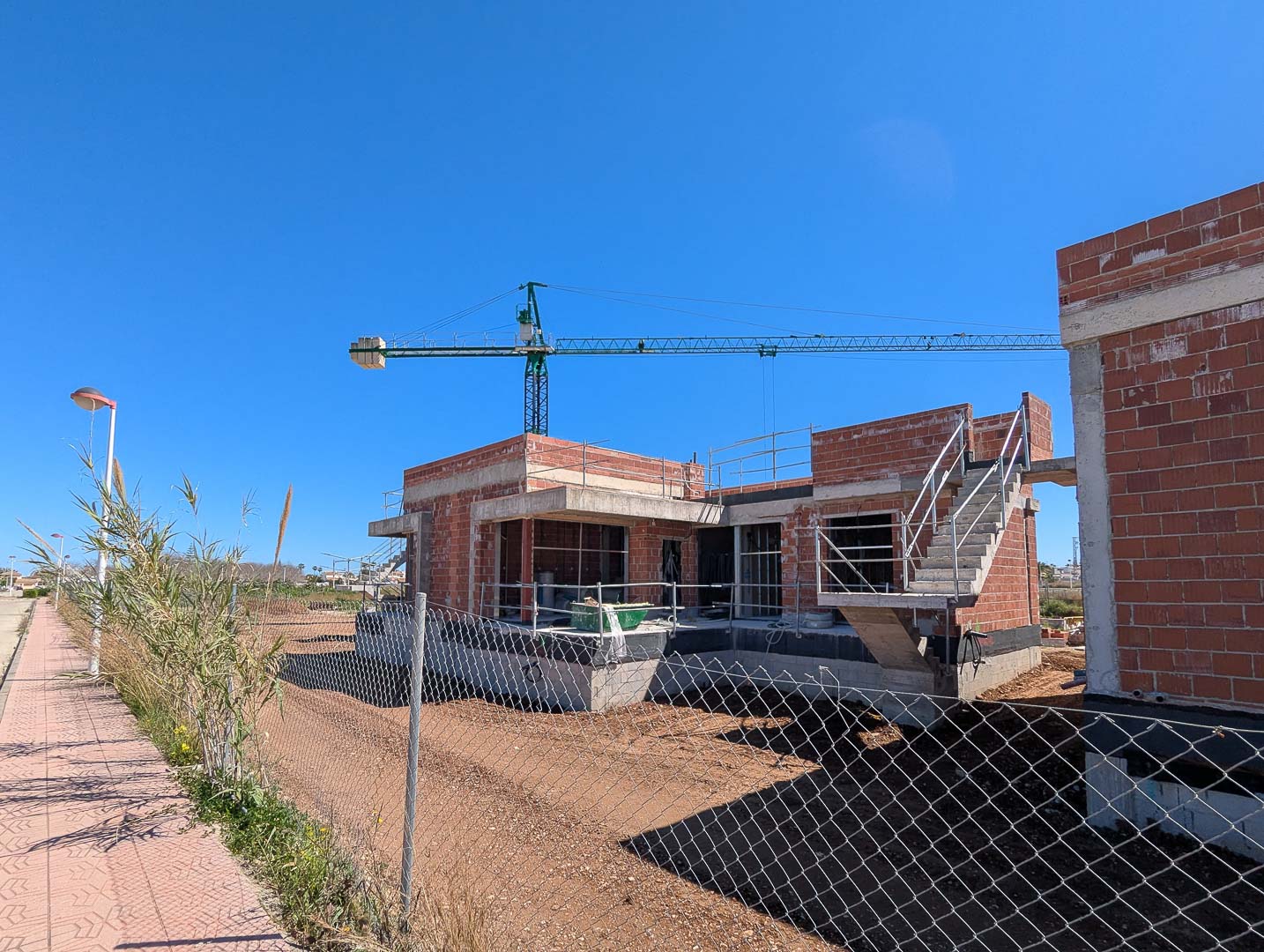 État actuel de la construction montrant la structure en briques rouges, les dalles en béton et l'escalier extérieur sur un terrain plat avec grue de chantier.