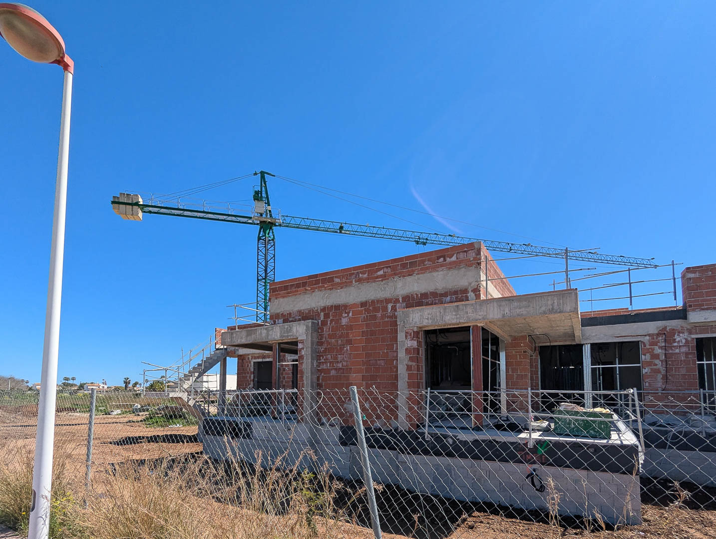 Phase actuelle de construction présentant la structure en briques et béton de la villa, avec grue de chantier et clôture sous un ciel bleu azur.