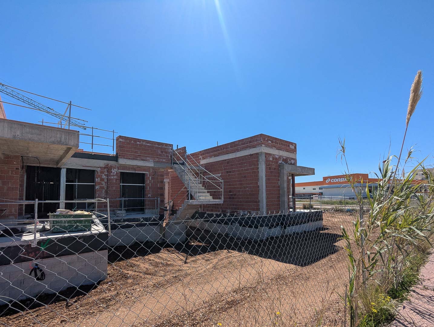 Vue extérieure de la villa en cours de construction, mettant en évidence la structure en briques, l'escalier en béton et la proximité des commerces.