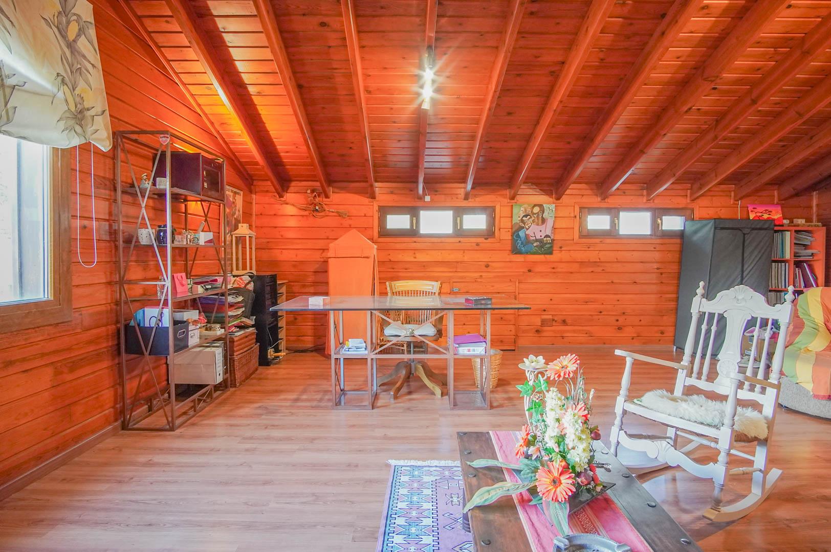 Bureau mansardé spacieux avec murs et plafonds en pin finlandais. L'espace comprend un bureau en verre et un coin lecture avec un fauteuil à bascule blanc traditionnel.