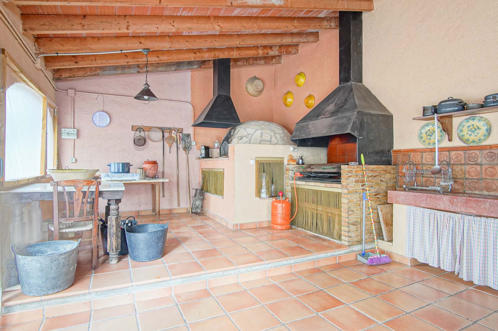 Cuisine d'été rustique avec four à bois traditionnel, barbecue en brique et charpente apparente en bois sur un sol en terre cuite.