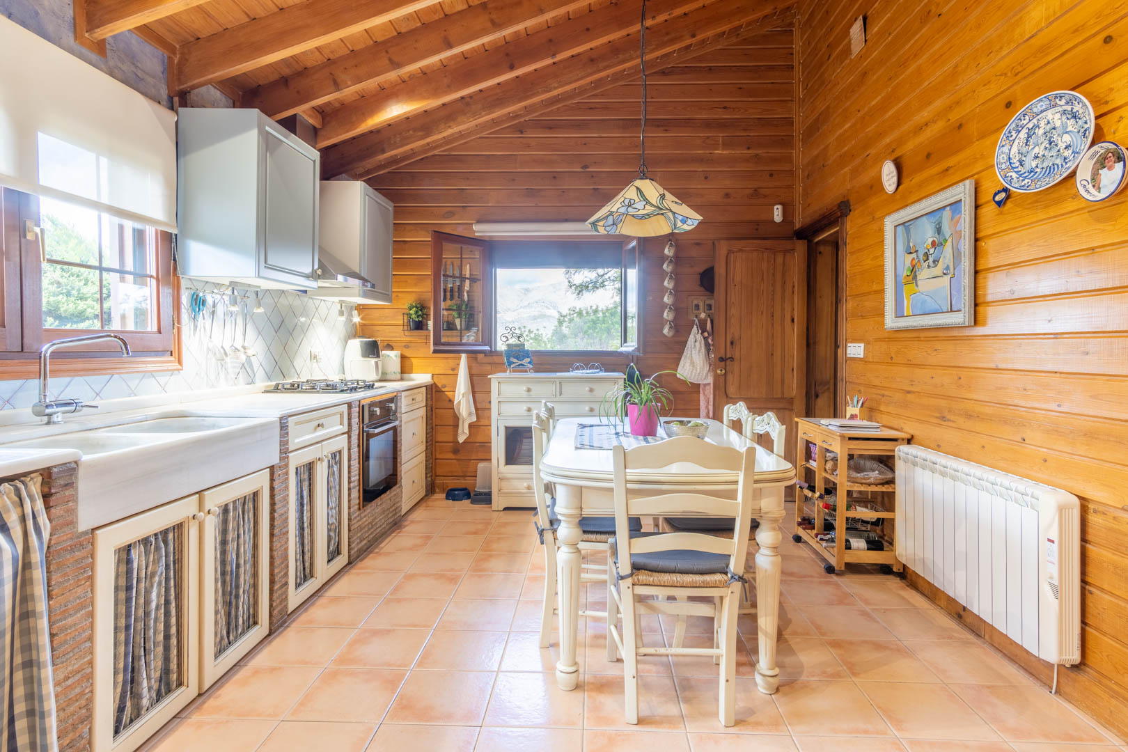 Cuisine rustique avec plafonds en pin finlandais, plan de travail en brique et pierre, et un coin repas central éclairé par une suspension de style Tiffany.
