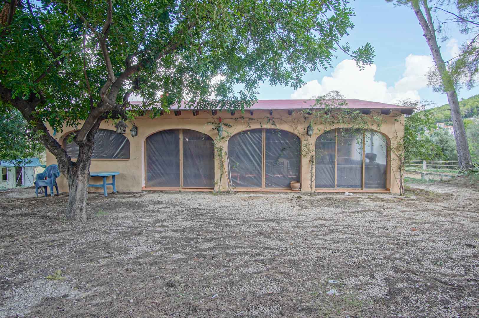Dépendance rustique avec des ouvertures en arche, des poutres apparentes et une façade aux tons naturels, située dans un espace extérieur arboré avec un sol en gravier.