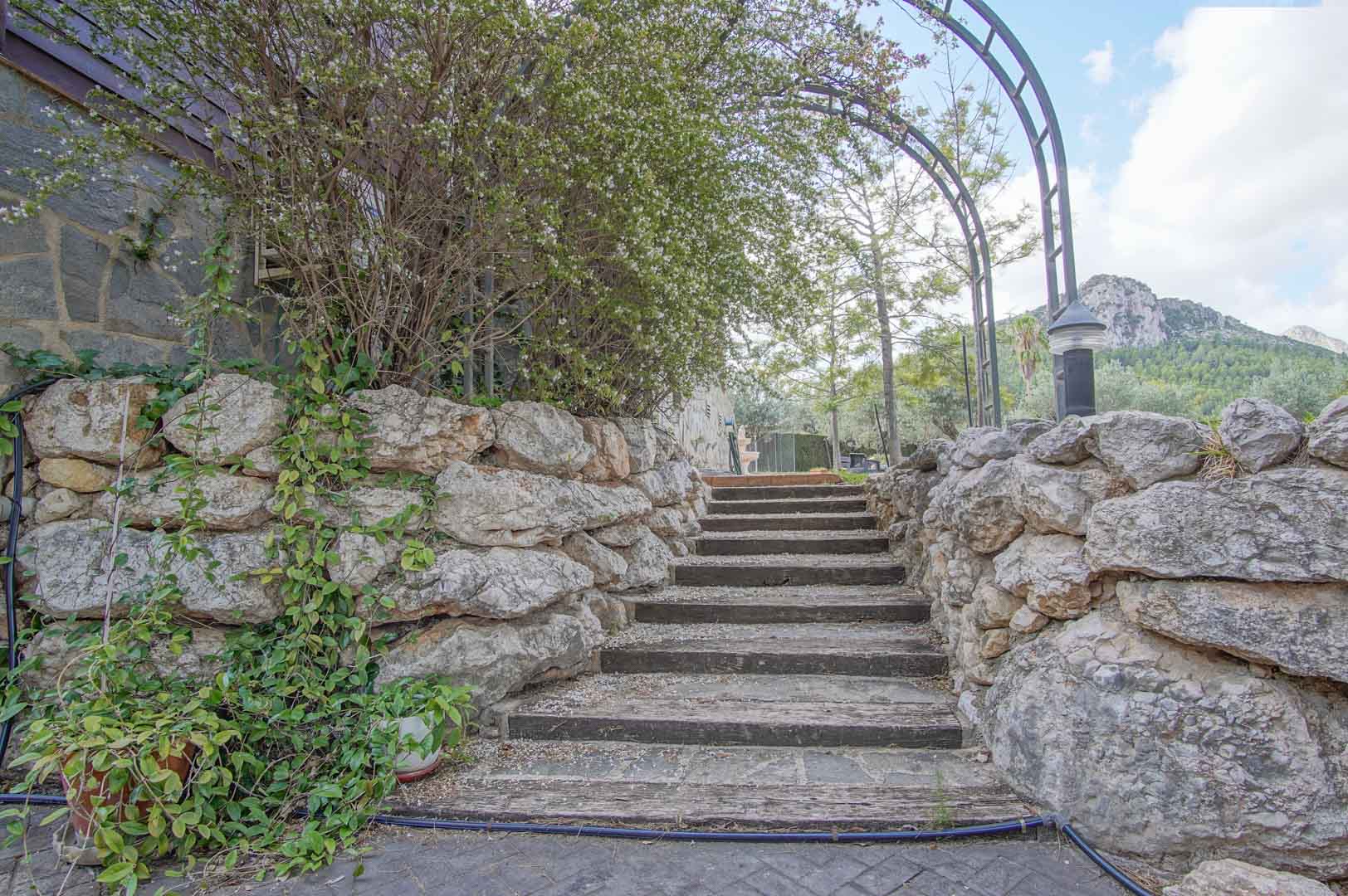 Escalier extérieur en traverses de bois et pierre naturelle, encadré par une arche métallique et des murets en pierre avec vue sur les montagnes.