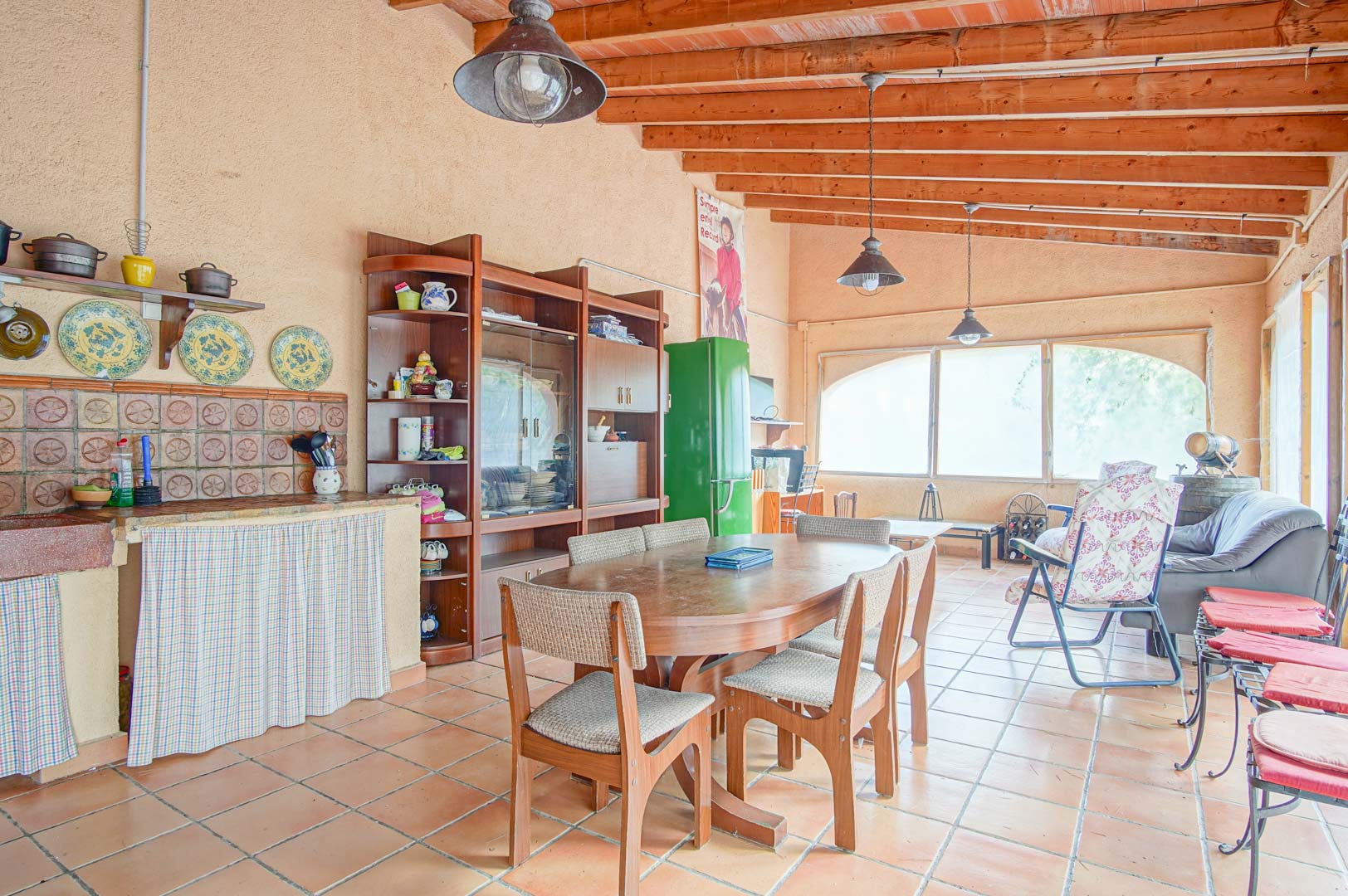 Espace de vie rustique avec poutres apparentes, sol en terre cuite, cuisine intégrée et coin repas lumineux sous un plafond en bois massif.