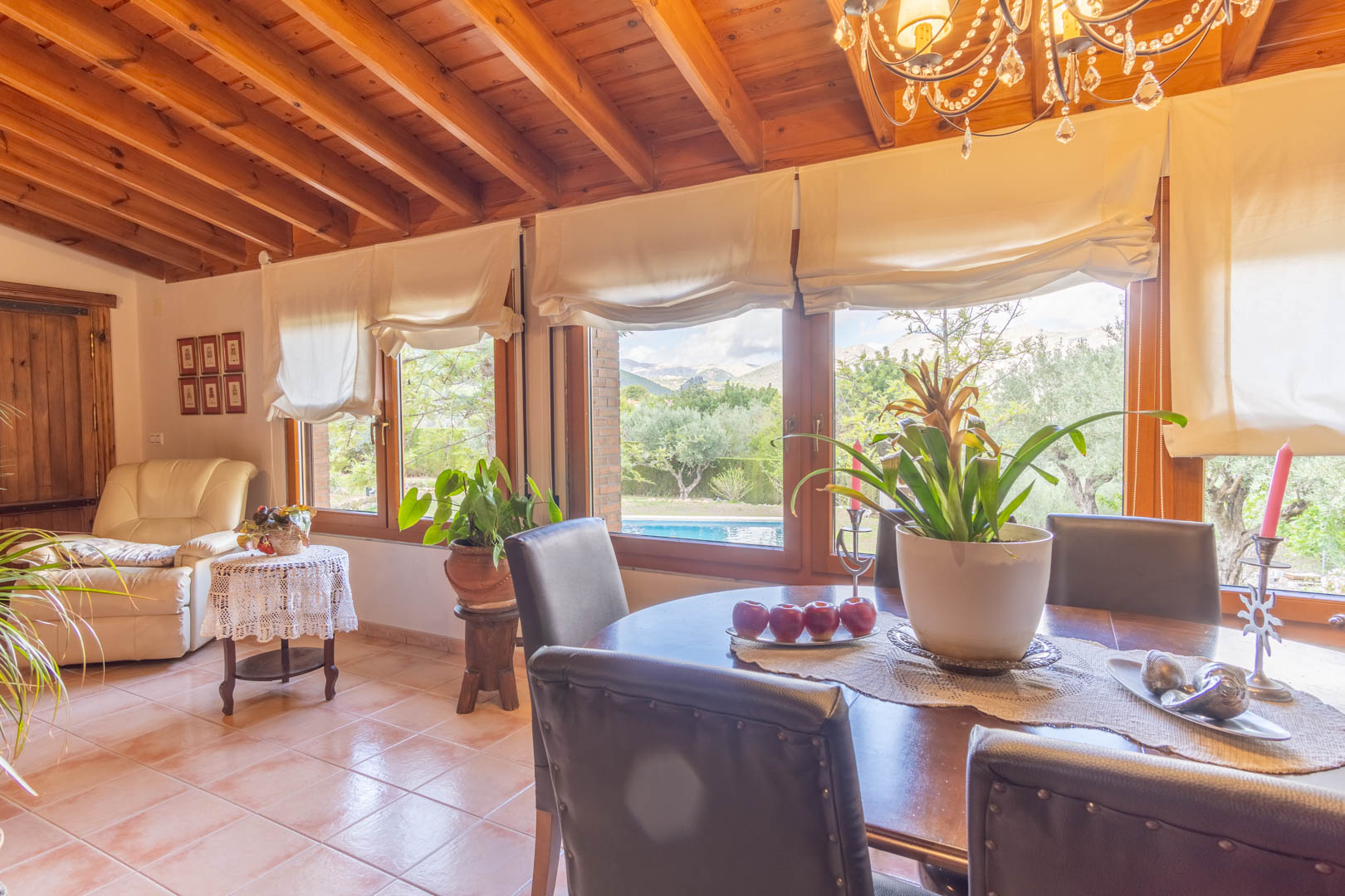 Salle à manger lumineuse avec plafond en poutres de pin finlandais, sol en terre cuite et grandes fenêtres offrant une vue sur la piscine et les montagnes d'Alicante.