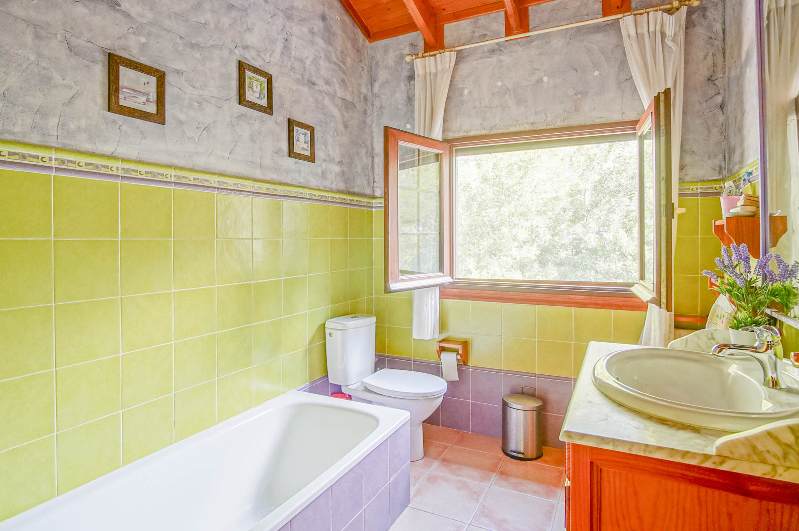 Salle de bain avec poutres en bois apparentes, carrelage vert et vasque sur plan en marbre. Une grande fenêtre s'ouvre sur la végétation environnante.