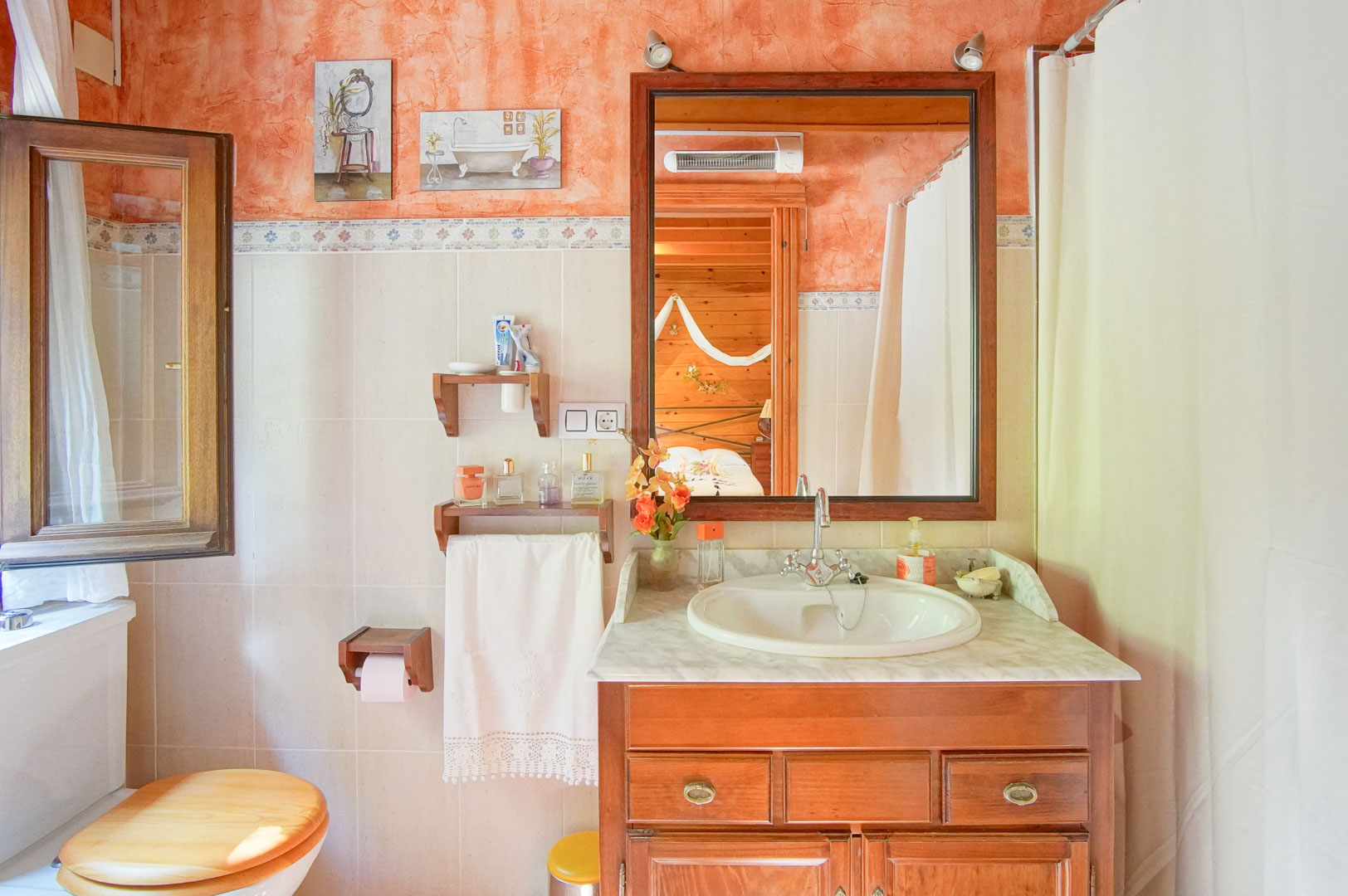Salle de bain rustique avec meuble sous-vasque en bois, plan en marbre et murs terre cuite, dont le miroir reflète la chambre adjacente aux finitions en pin.