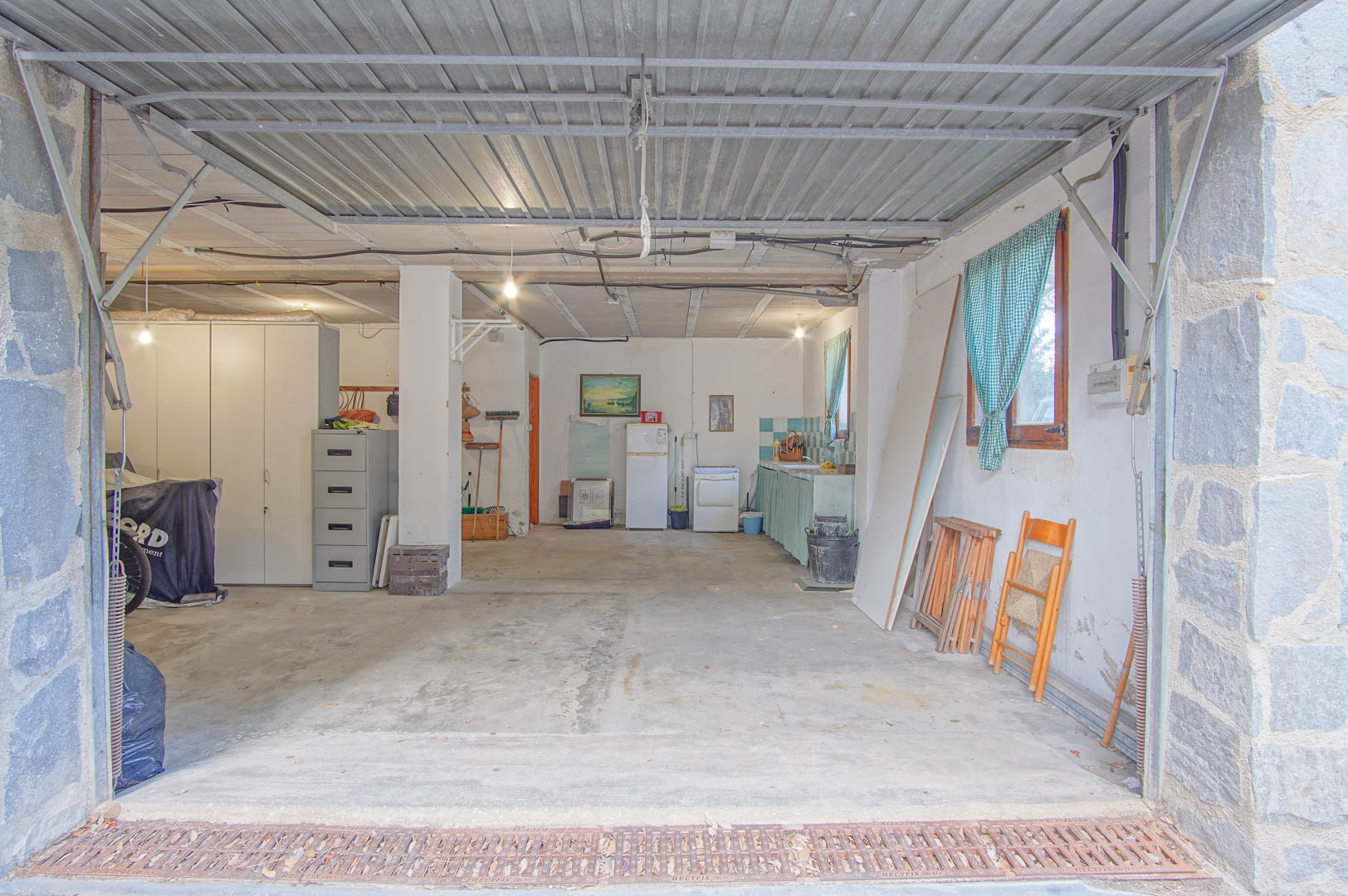 Vaste demi-sous-sol comprenant un garage pour plusieurs véhicules, un atelier et une kitchenette. L'espace dispose de sols en béton, de piliers en pierre et d'une porte métallique.