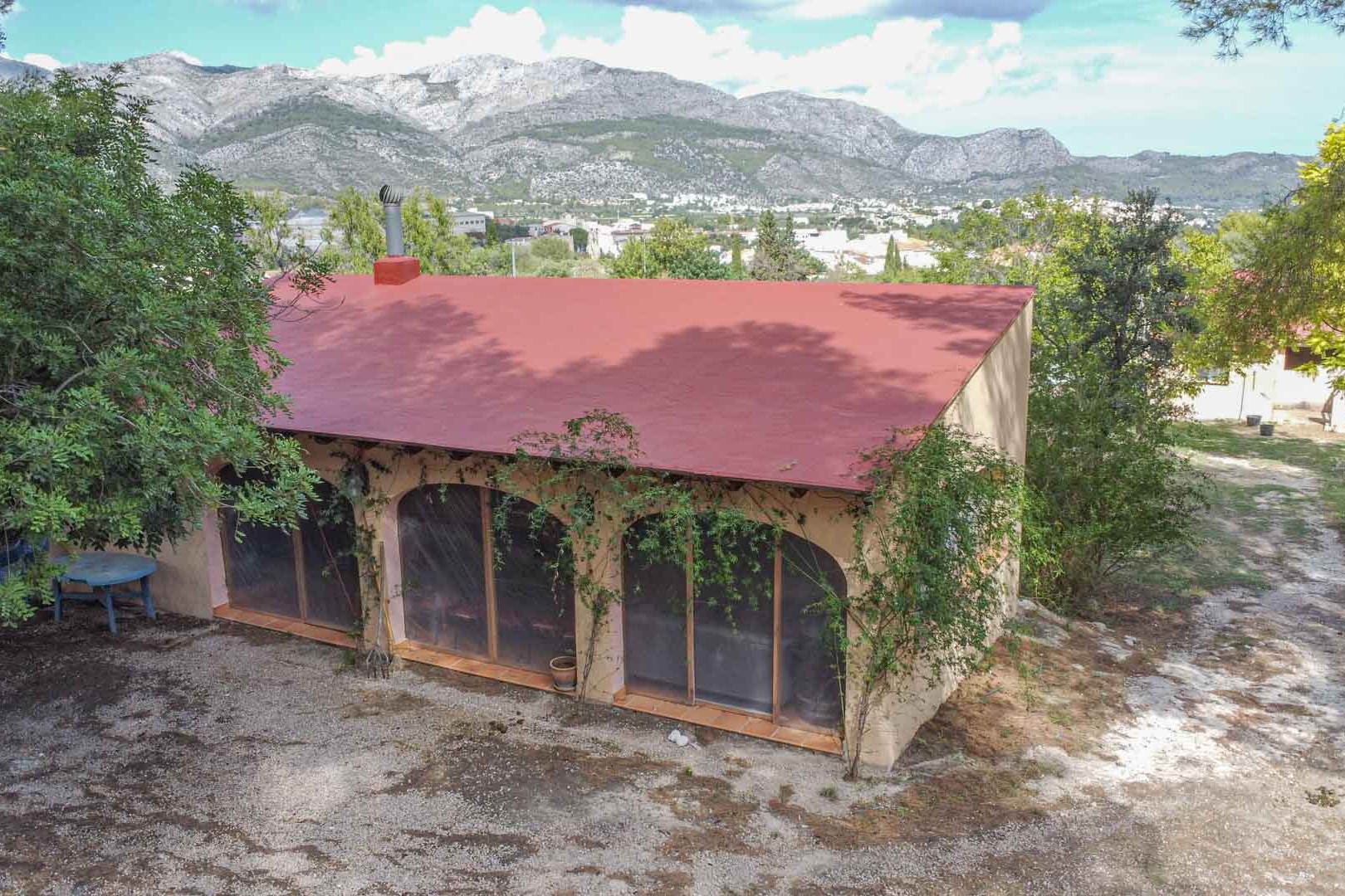 Vue extérieure de la dépendance avec ses grandes arches vitrées et sa cheminée, située dans un environnement naturel offrant une vue dégagée sur les montagnes.