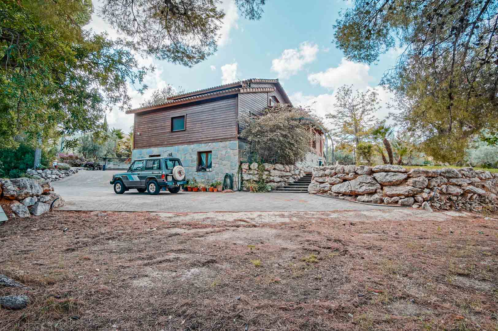 Vue extérieure de la villa avec façade en pierre naturelle et bois de pin, une large allée pavée et des murs de soutènement en pierre sèche.
