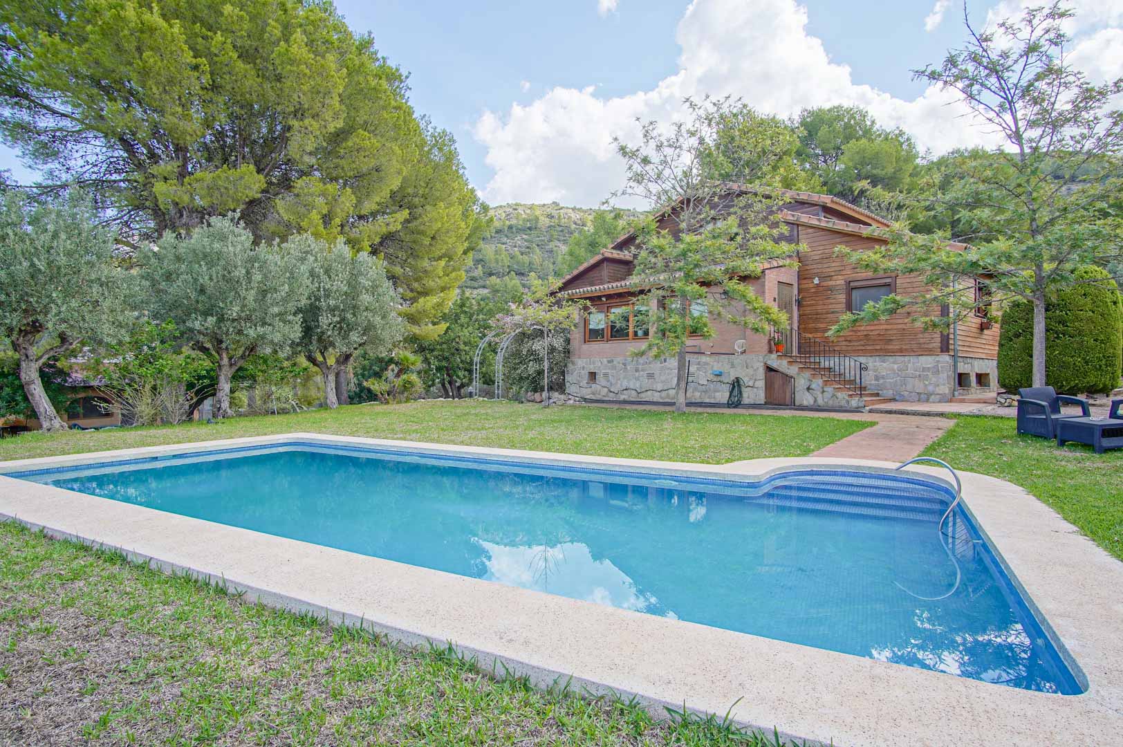 Vue extérieure de la villa avec sa piscine rectangulaire, mettant en valeur la façade en pin finlandais et pierre naturelle au cœur d'un jardin méditerranéen boisé.