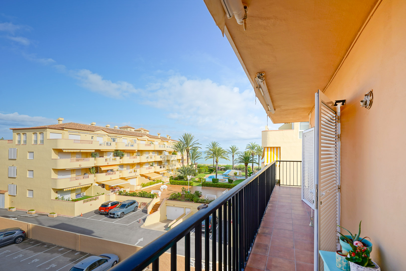 Balcon avec carrelage en terre cuite et garde-corps noir offrant une vue sur la piscine commune, les jardins et l'horizon marin.