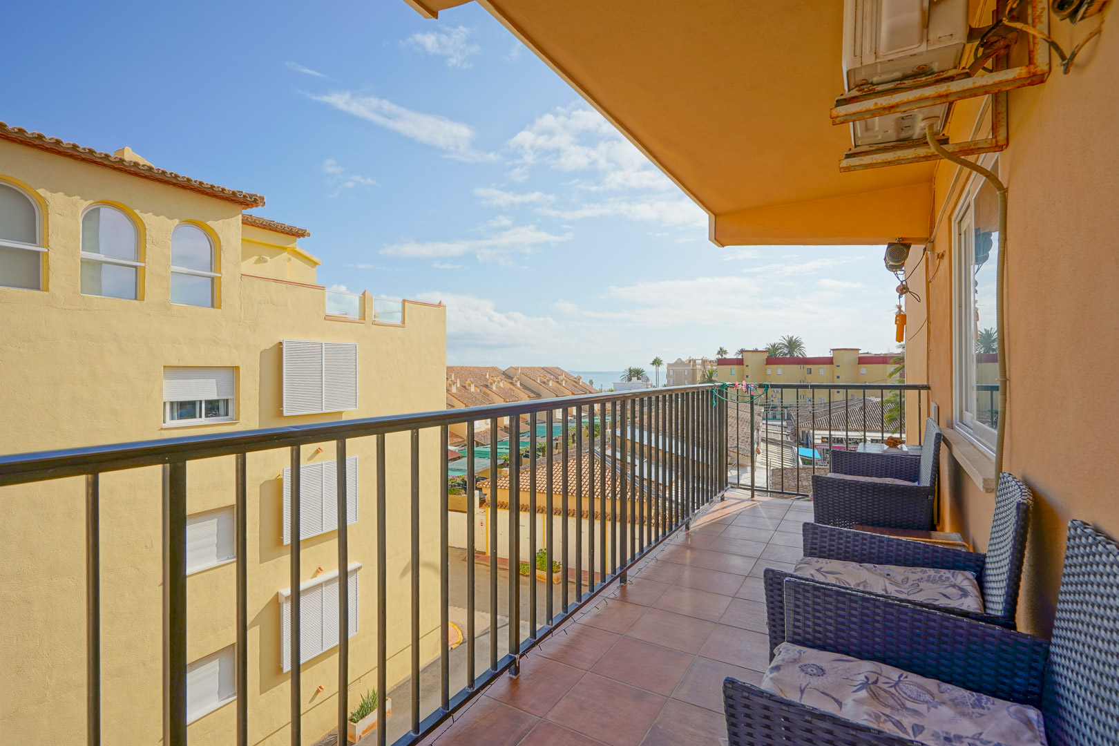 Balcon privé avec sol en terre cuite, garde-corps en métal noir et fauteuils en rotin. Il offre une vue sur les bâtiments environnants et l'horizon marin au loin.